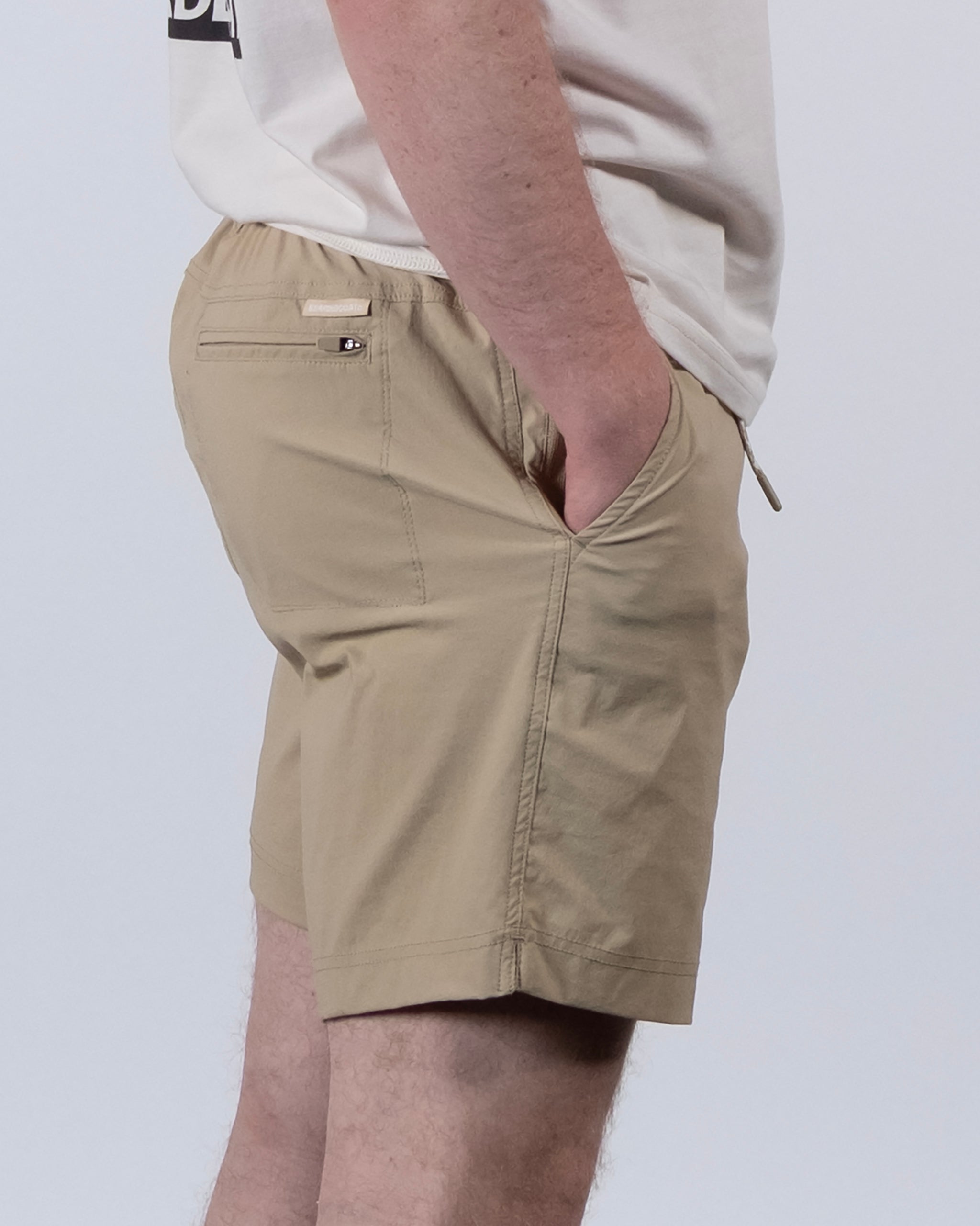 A man in a white shirt and BEARDEDGOAT Men's Rover Hybrid Short in light khaki stands sideways with hands in pockets against a plain white background.