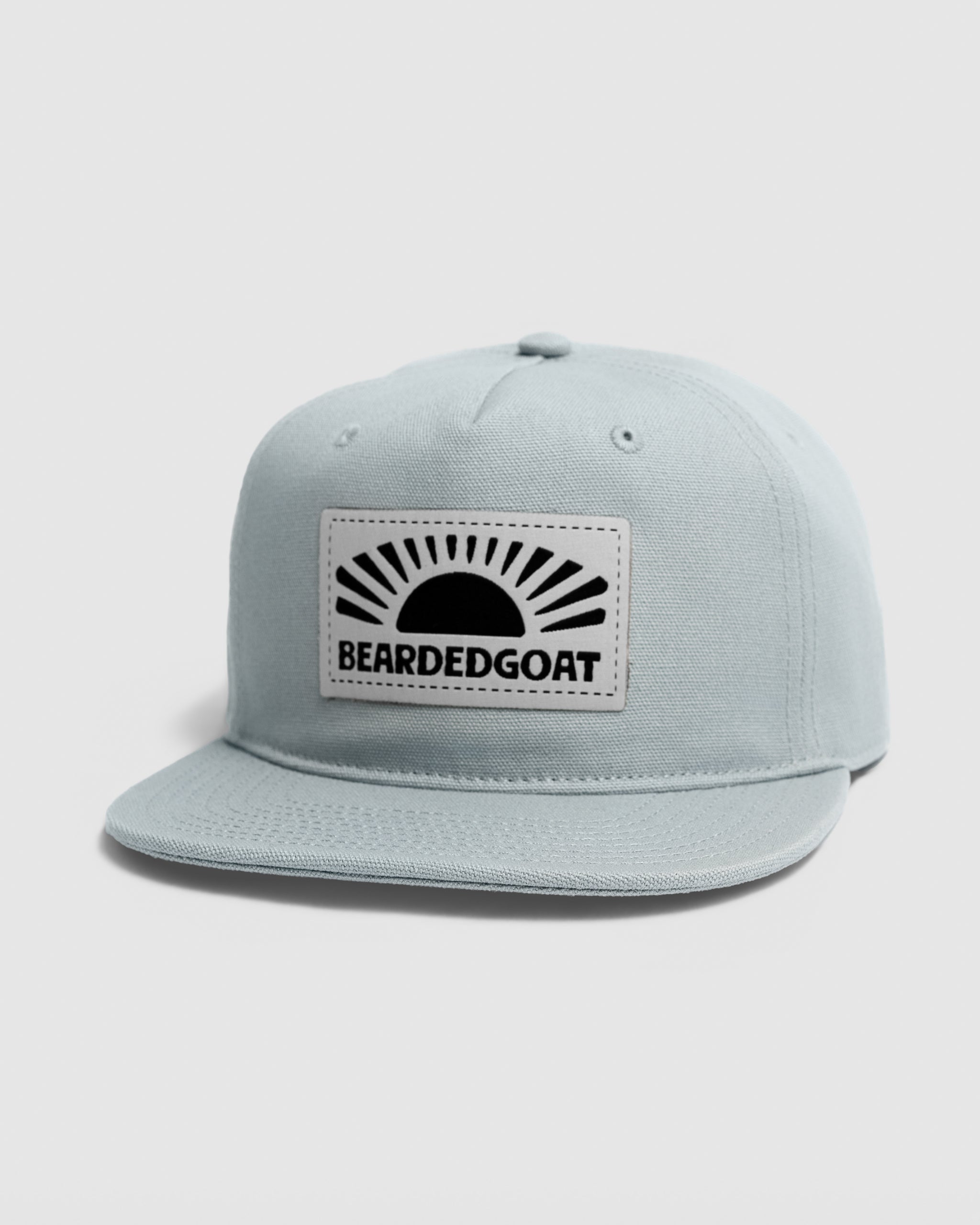 The BEARDEDGOAT Sunrise Label Hat - Grey features a flat brim and a front rectangular patch with a black sunburst and bold BEARDEDGOAT text—ideal for outdoor style and adventure.