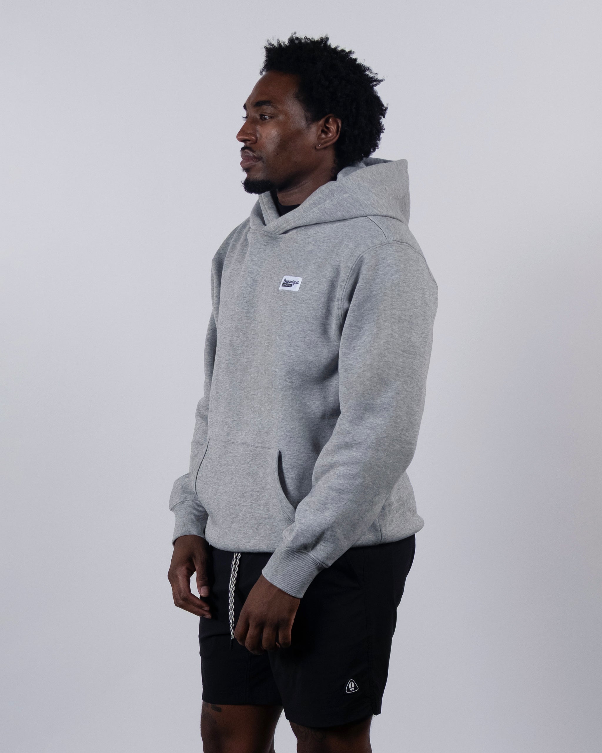 A man stands against a light gray background, wearing the beardedgoat VOID™ Hoodie—crafted from heavyweight cotton fleece with a small white chest patch—and black shorts. He looks left, hands relaxed at his sides.
