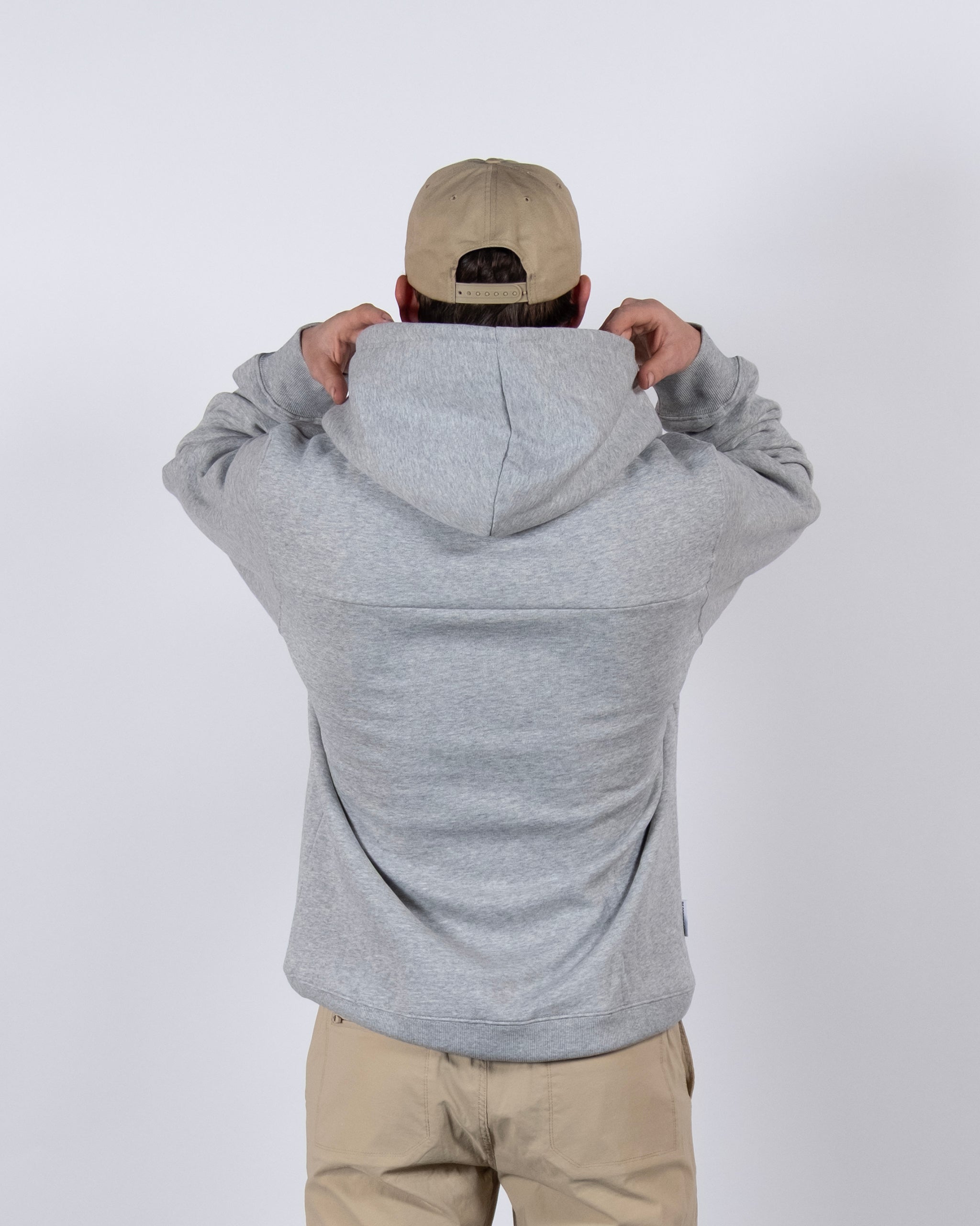 A person faces away, holding up the hood of a beardedgoat The VOID™ Hoodie in premium heavyweight cotton (light gray), paired with beige pants and a beige baseball cap, set against a plain white background.