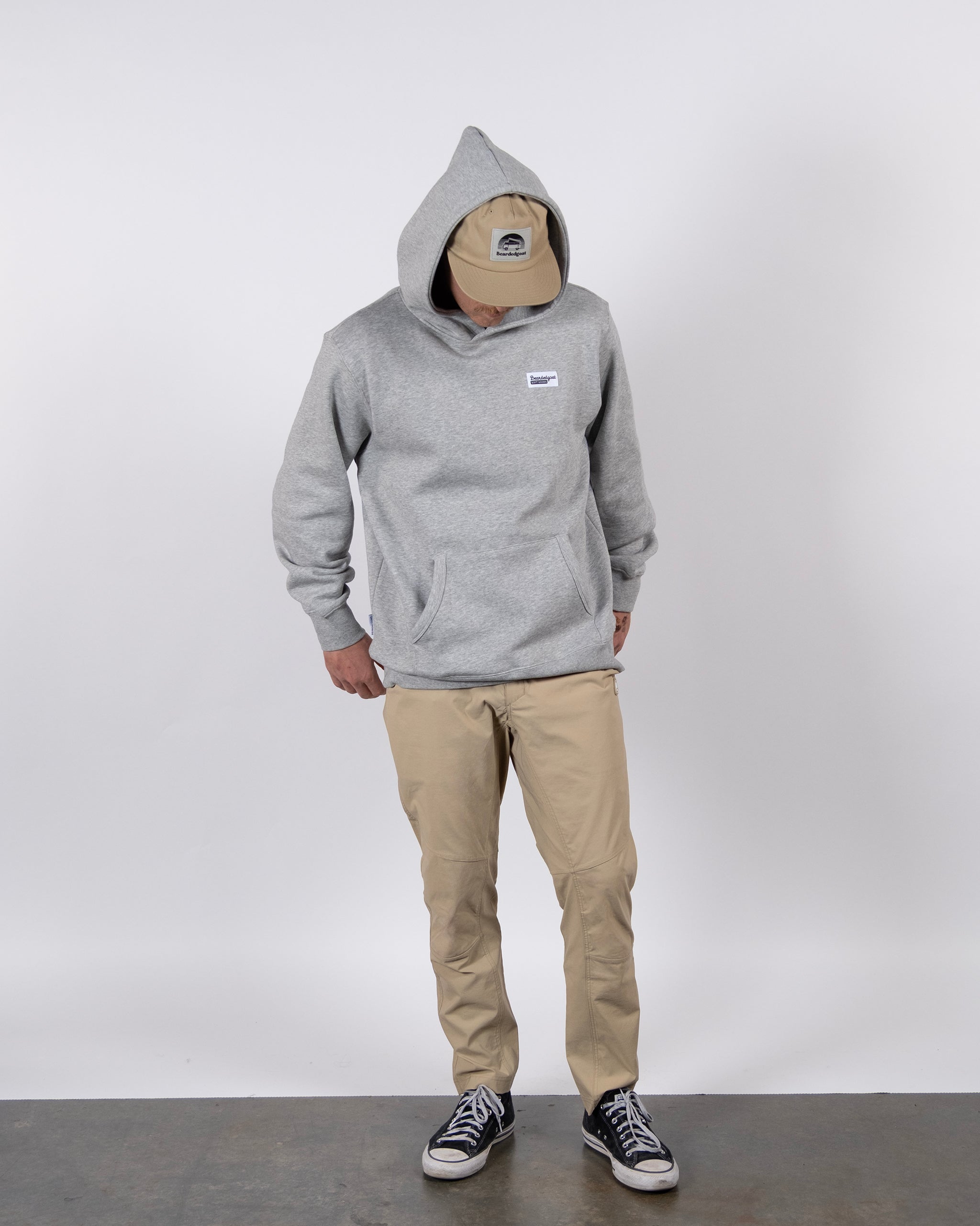 Wearing the beardedgoat VOID™ Hoodie in grey with the hood up, a beige cap, khaki pants, and black sneakers, a person stands against a white background looking down with hands in the hoodie pockets.