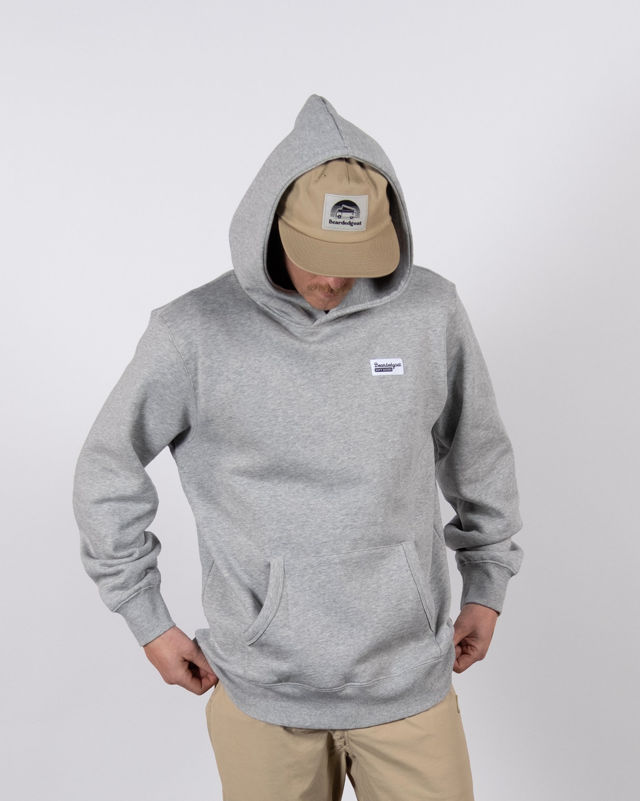 A person wearing the beardedgoat The VOID™ Hoodie in gray, featuring premium heavyweight cotton, a front pocket, and a small white label, pairs it with a beige cap and pants while standing against a plain white background.