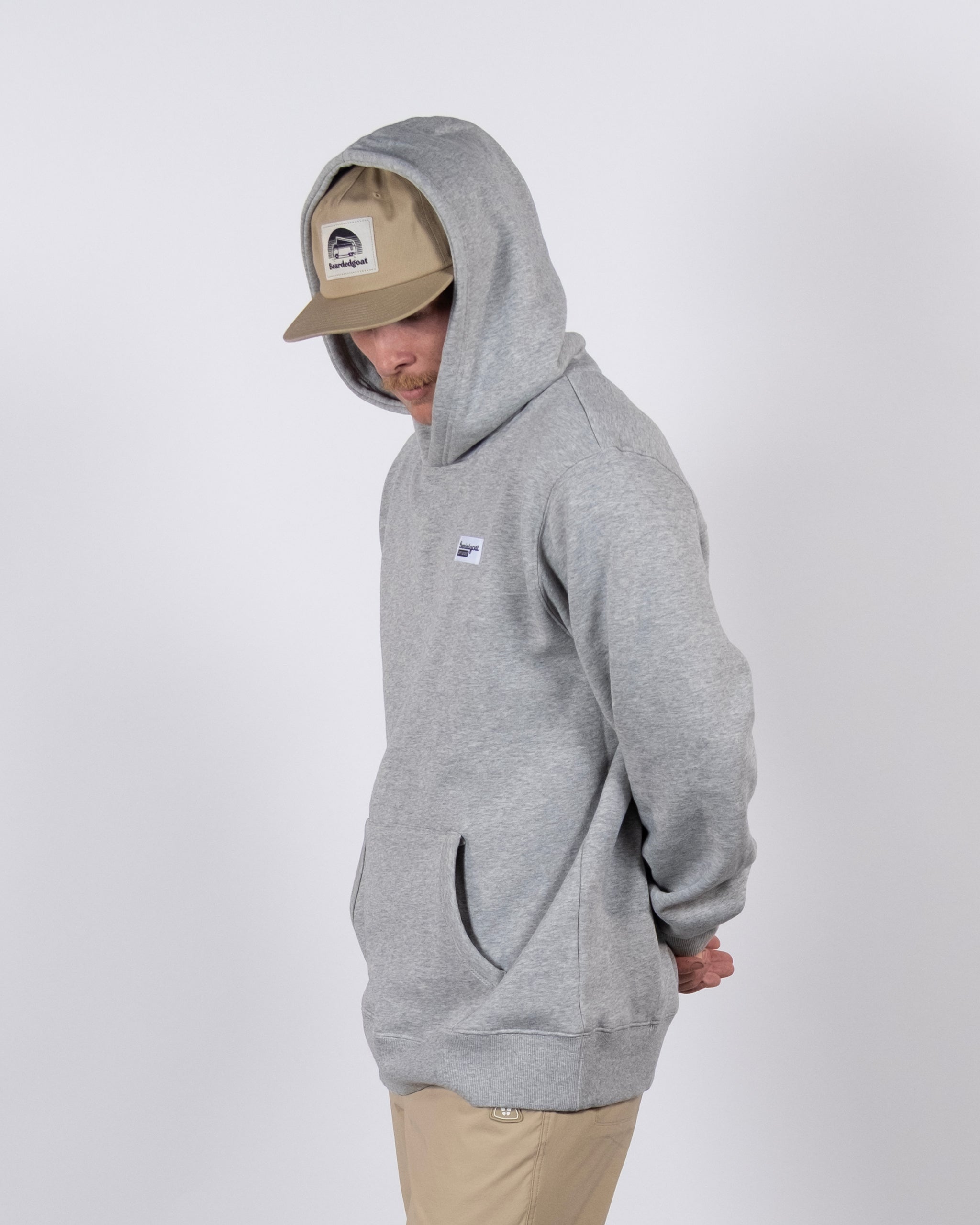 A person wearing the beardedgoat The VOID™ Hoodie in premium heavyweight cotton, light gray color, with the hood up and a beige cap, stands against a plain white background, facing slightly downward and left with hands behind their back.