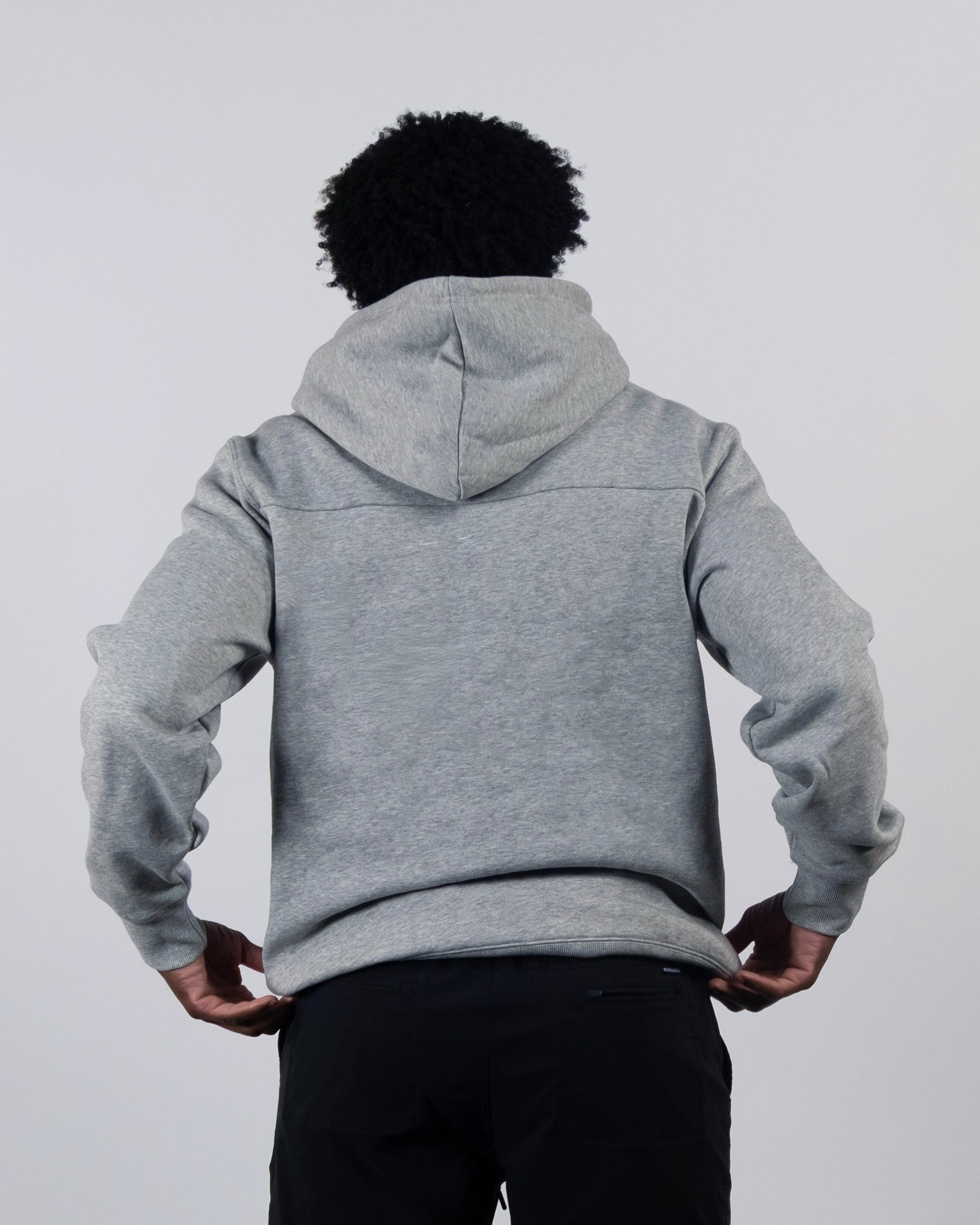 A person with curly hair, dressed in black pants and a beardedgoat The VOID™ Hoodie in gray heavyweight cotton fleece, stands with their back to the camera against a plain light background.