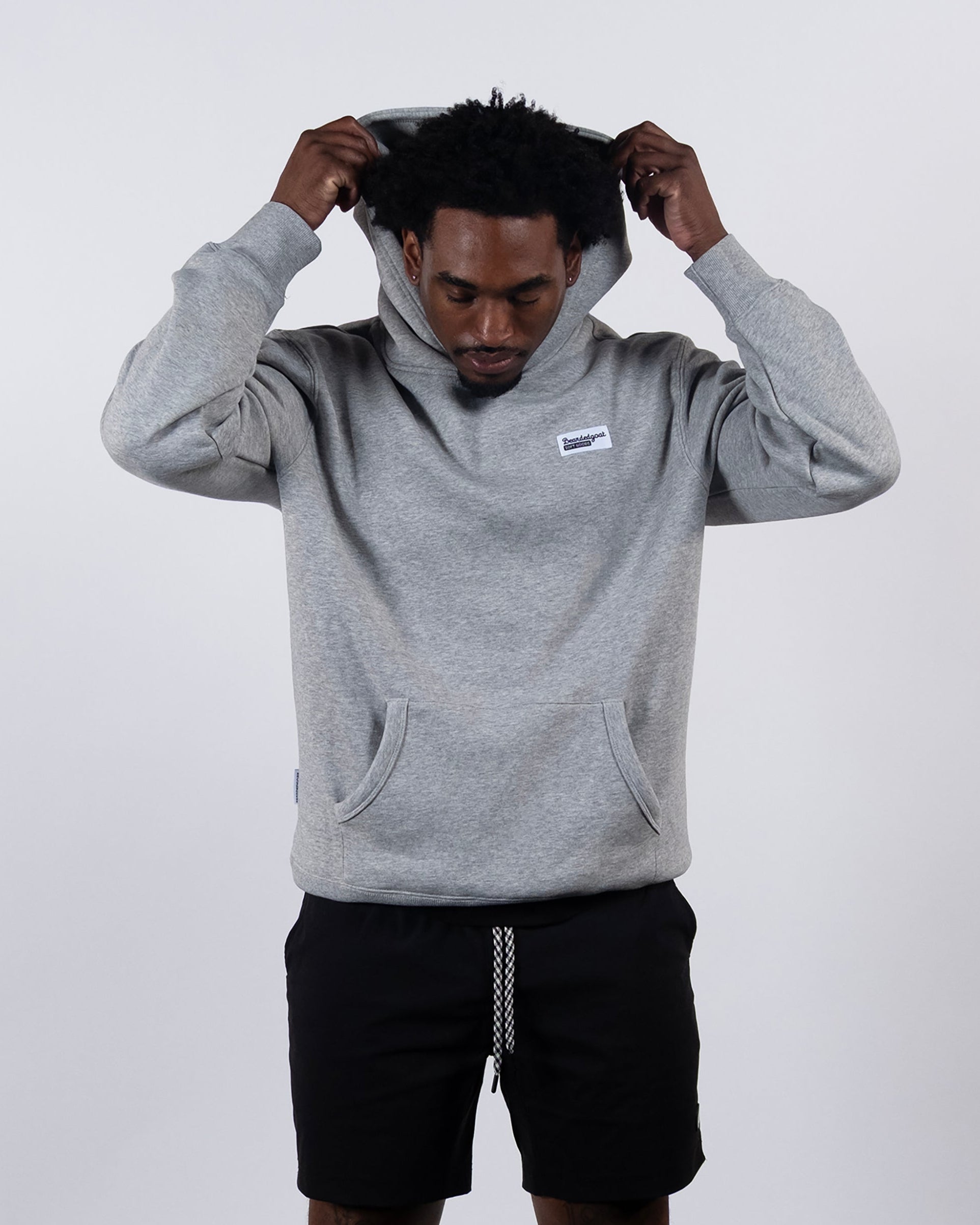 A man faces forward against a plain white background, pulling the hood over his head while wearing the beardedgoat The VOID™ Hoodie in gray, featuring heavyweight cotton fleece, a small white label, and black drawstring shorts.