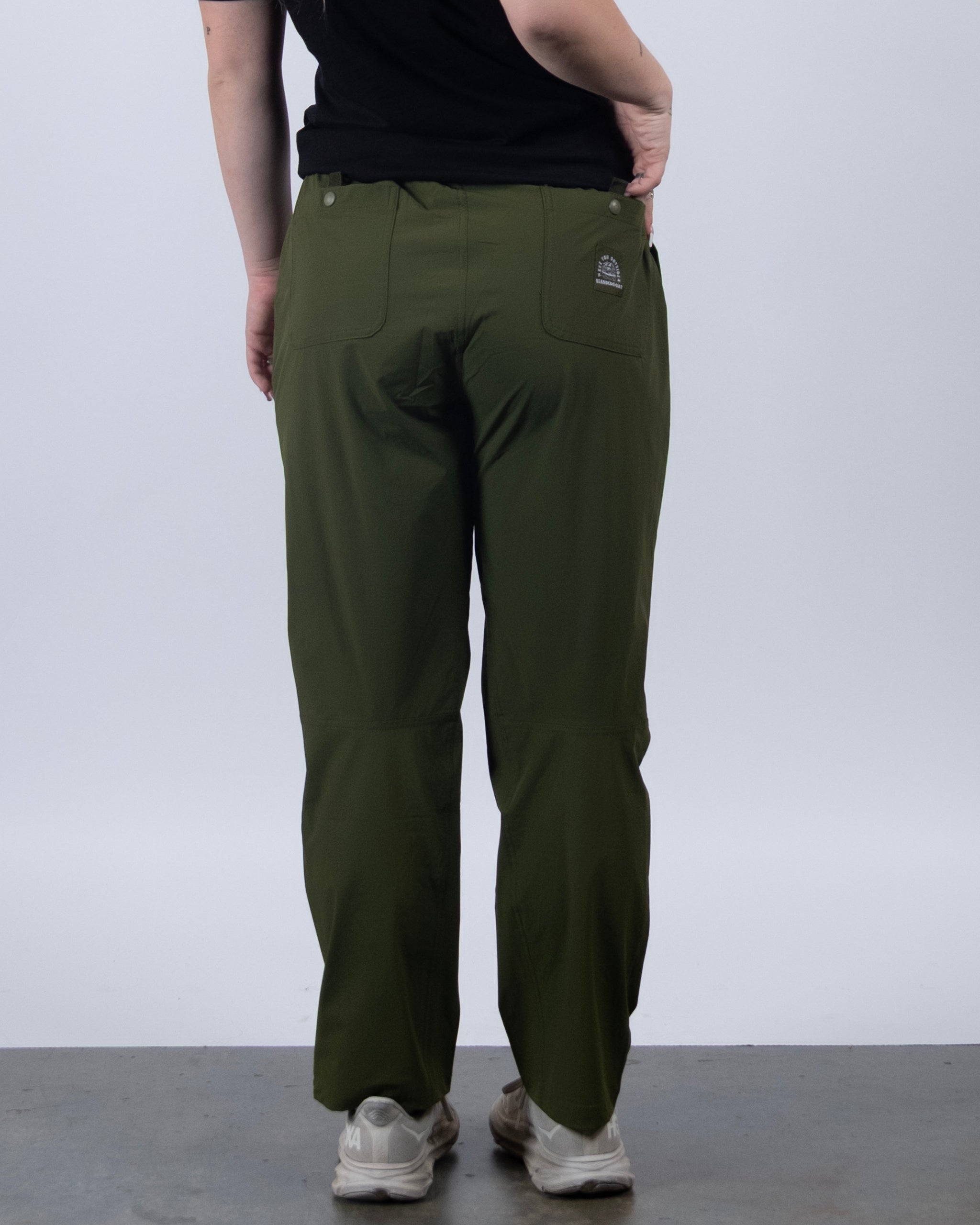 A person wearing BEARDEDGOAT’s NEW — Summit™ Pant in olive green and white sneakers stands with their back to the camera against a plain light background, resting their left hand on their hip.