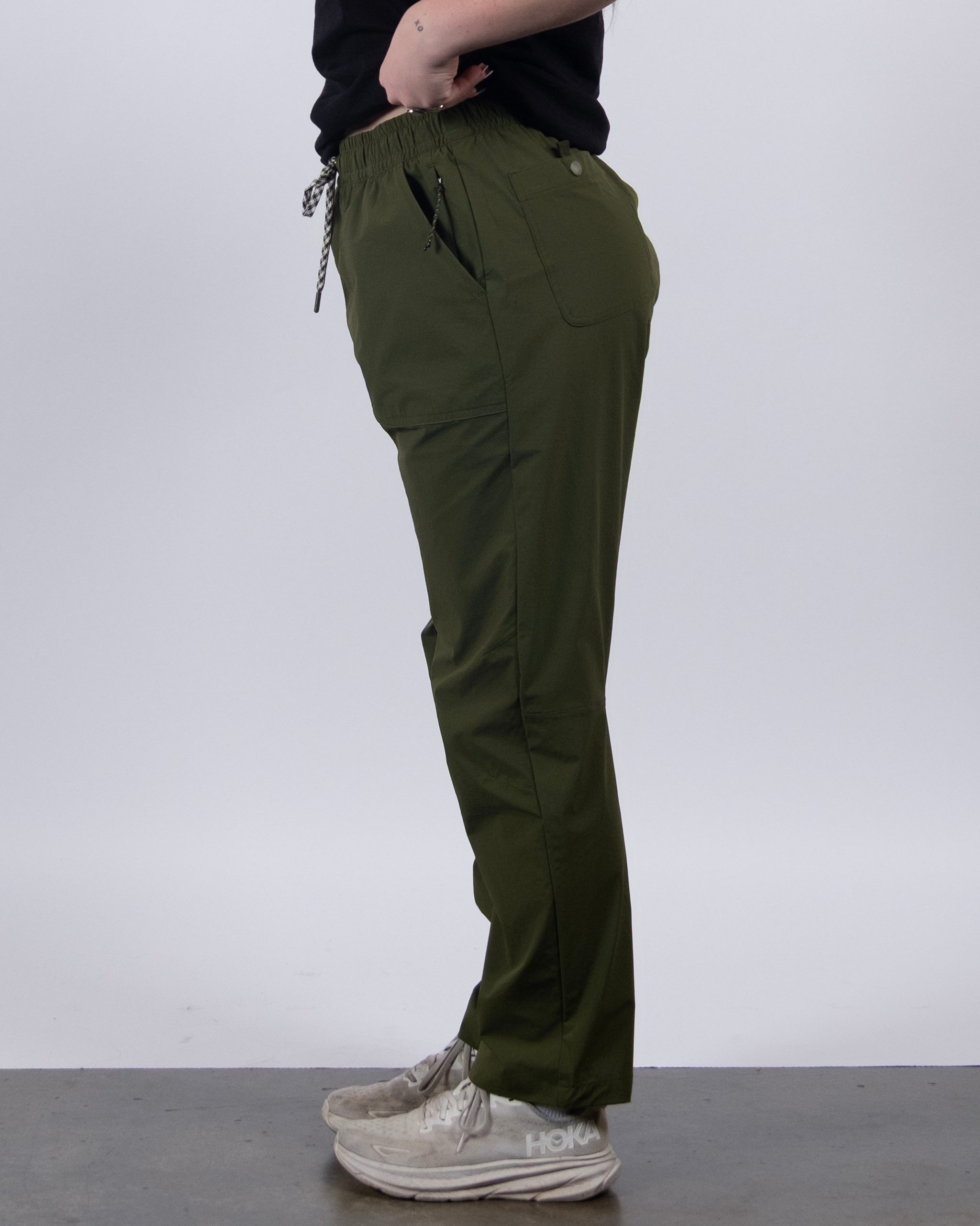 A person in profile wears BEARDEDGOAT’s NEW — Summit™ Pant in olive green, featuring an elastic waistband with drawstring and side pockets, paired with a black shirt and white sneakers against a plain light gray background.