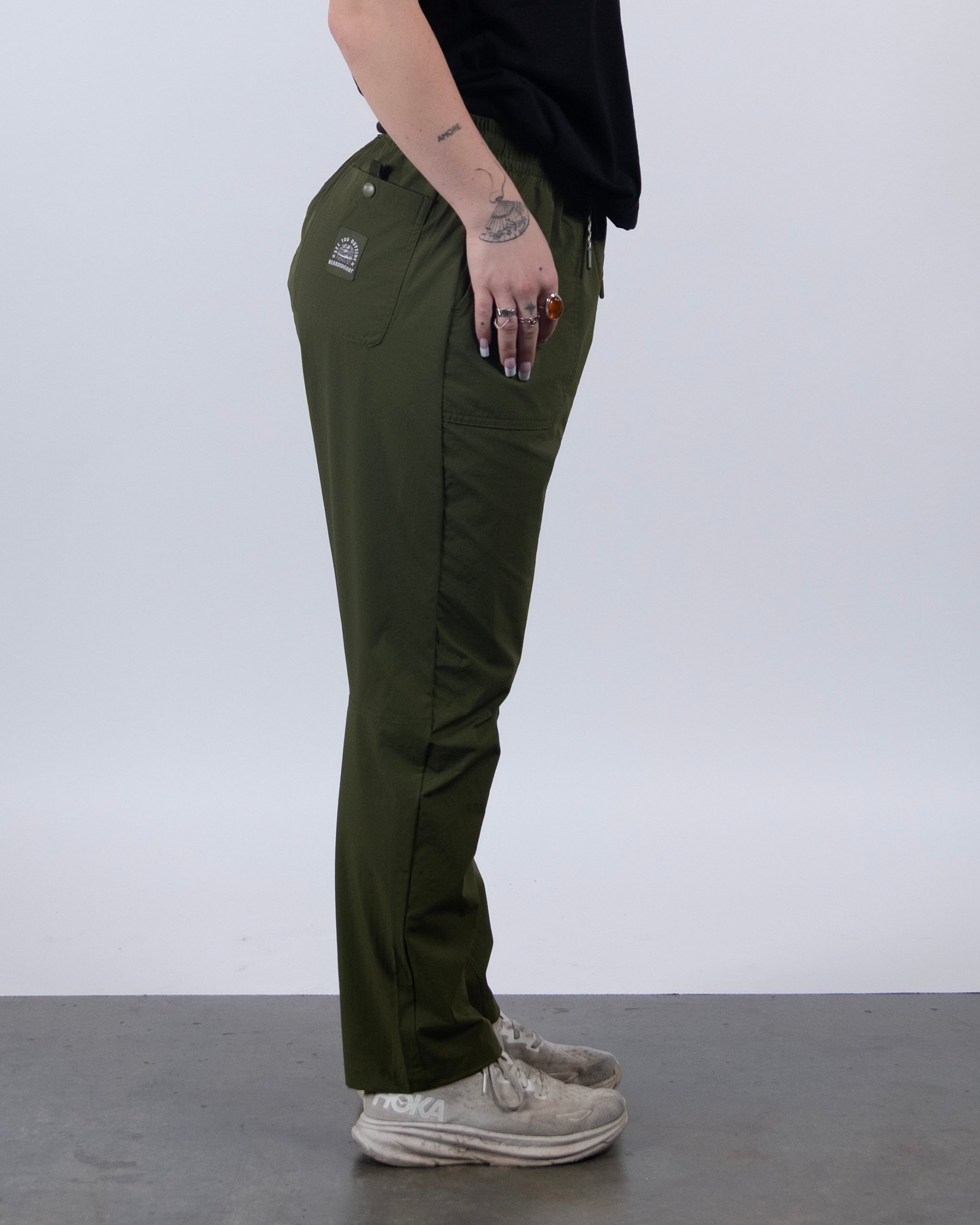 A person stands sideways against a plain light background, wearing BEARDEDGOAT’s NEW — Summit™ Pant in olive green, a black shirt, white sneakers, rings, and showing tattoos plus visible pocket logos on the stain-resistant pants.