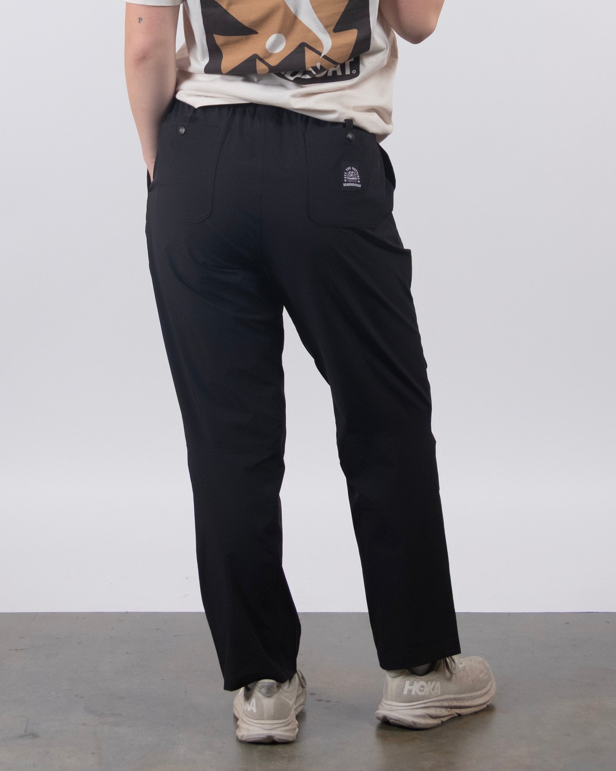 A person stands on a plain white floor seen from behind, wearing a white graphic T-shirt, BEARDEDGOAT NEW — Summit™ Pant with a logo on the back pocket, and light gray Hoka sneakers. The background is plain white.