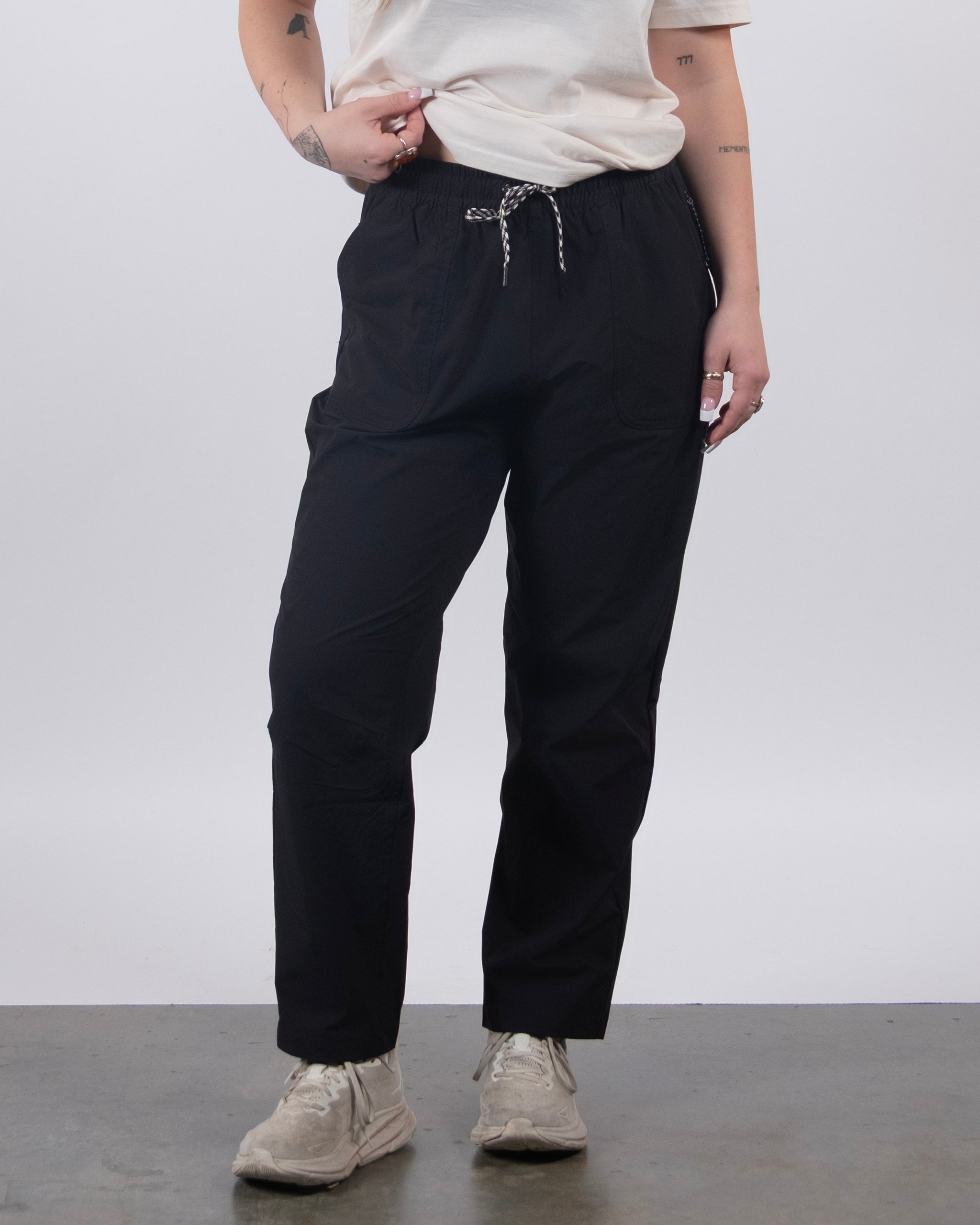A person wears the BEARDEDGOAT NEW — Summit™ Pant: black drawstring performance pants styled with a white t-shirt and sneakers. They hold the waistband, displaying arm tattoos, against a plain gray background.