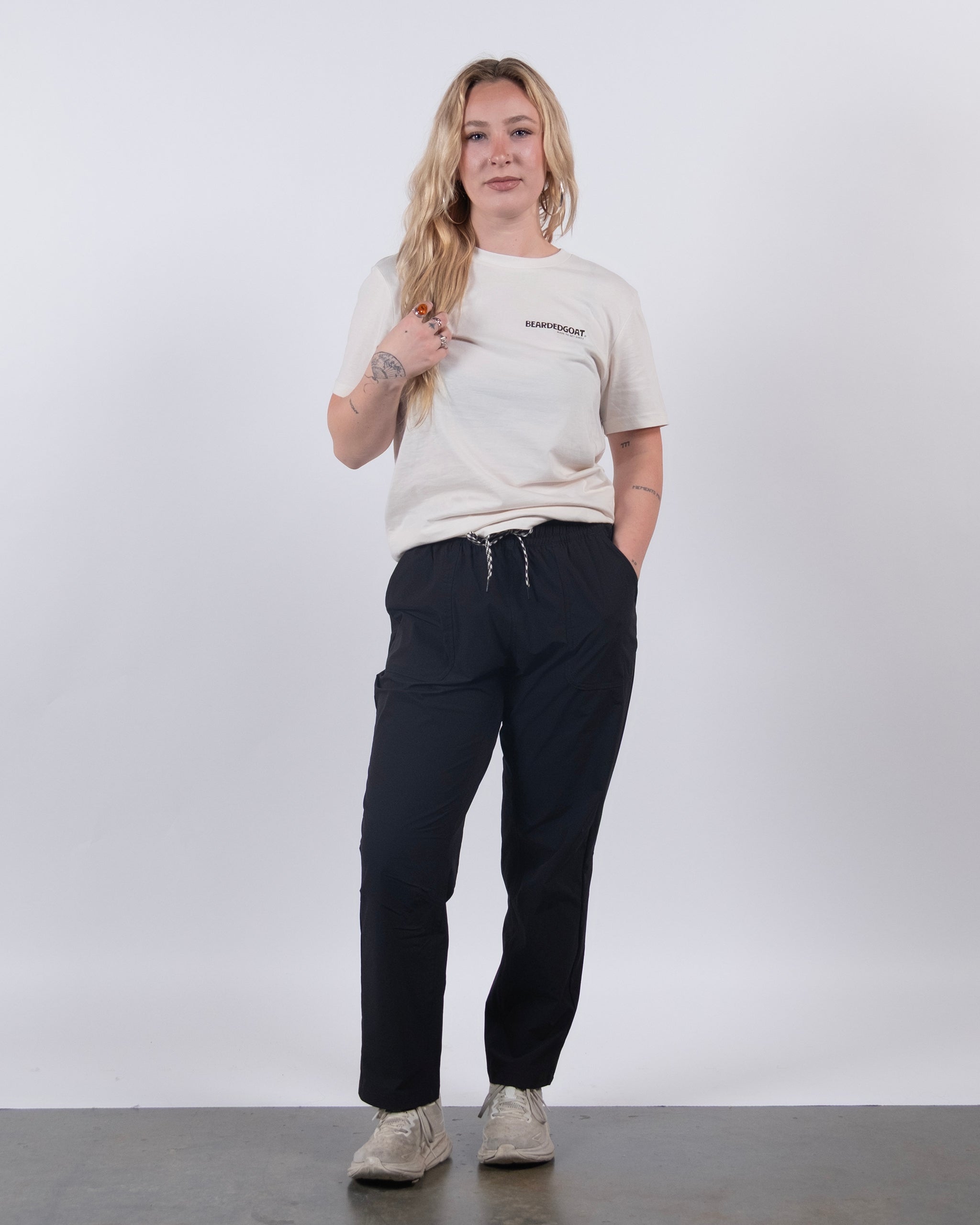 A person with long blonde hair stands against a white background, wearing the BEARDEDGOAT NEW — Summit™ Pant, a light T-shirt, and light sneakers. One hand is in their pocket while the other touches their hair as they smile slightly.