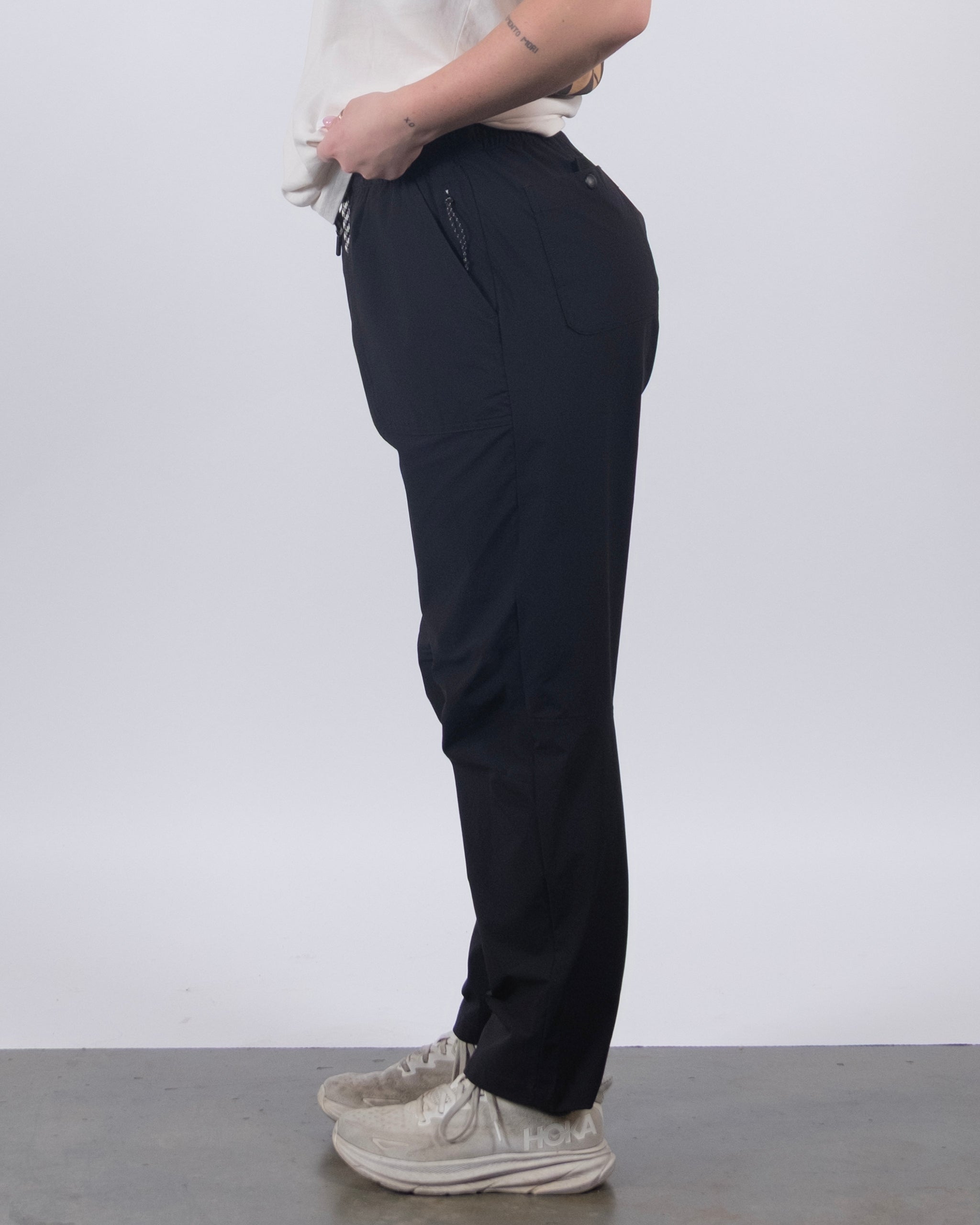 A person stands sideways against a plain background, wearing a white shirt, white sneakers, and BEARDEDGOAT’s NEW — Summit™ Pant: stain-resistant women’s performance pants with zippered pockets.