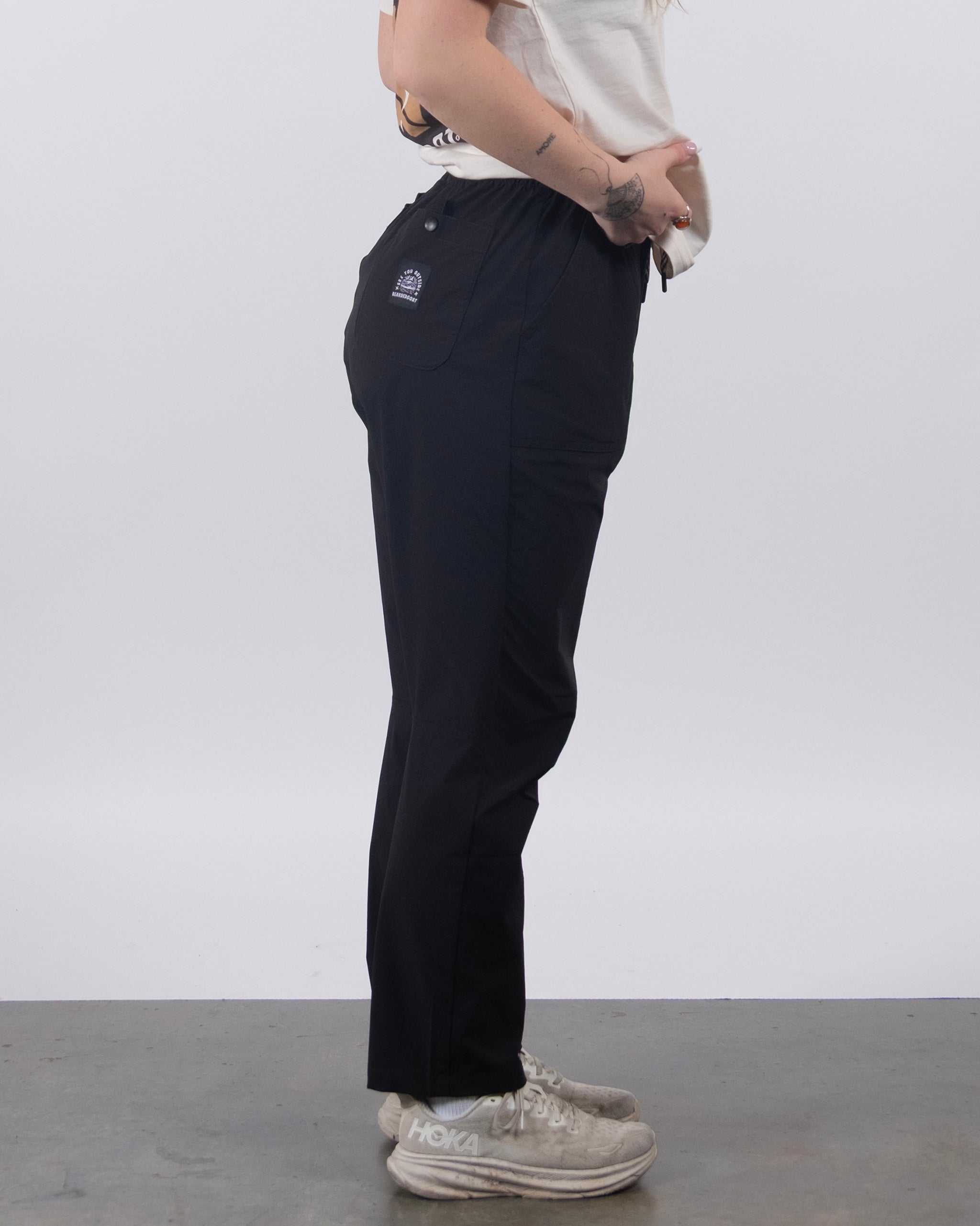 A person models BEARDEDGOAT's NEW — Summit™ Pant with a white t-shirt and sneakers, standing sideways with hands on hips. The background is plain white with a gray floor.