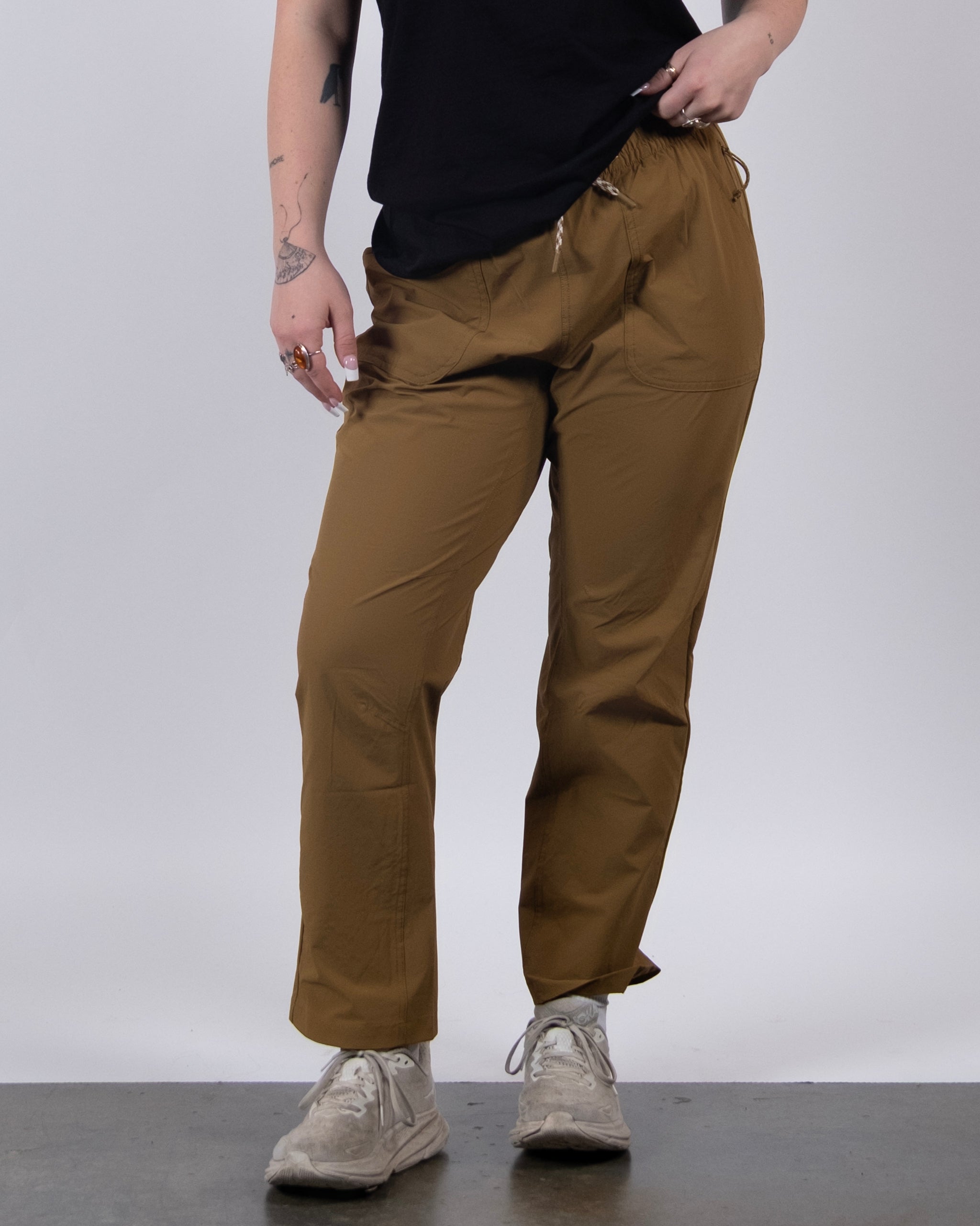 A person is wearing BEARDEDGOAT’s NEW — Summit™ Pant in brown with a black t-shirt and white sneakers, standing against a plain light background. Only their lower body and arms are shown.
