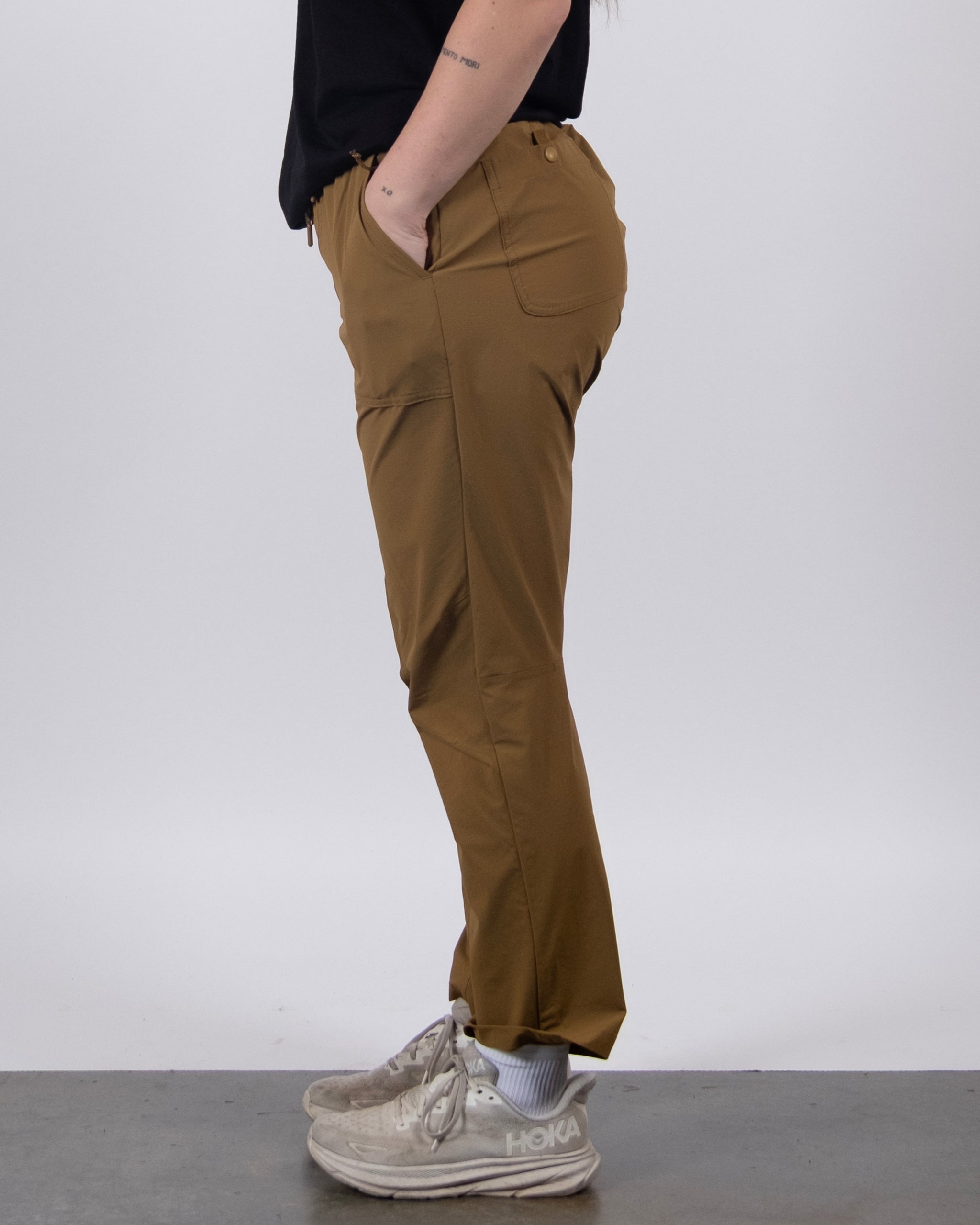 Only the lower half of a person is visible as they stand in profile against a plain background, wearing a black top, BEARDEDGOAT NEW — Summit™ Pant with one hand in the pocket, and white Hoka sneakers.