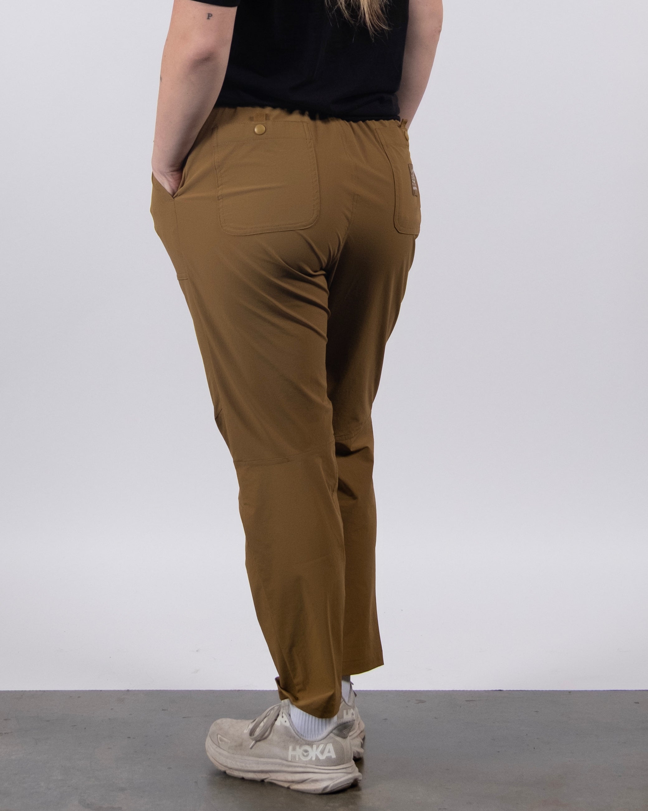Someone wearing BEARDEDGOAT's NEW — Summit™ Pant in brown, a black shirt, and white Hoka sneakers stands on a gray floor with their back to the camera and hands in pockets, set against a plain white background.