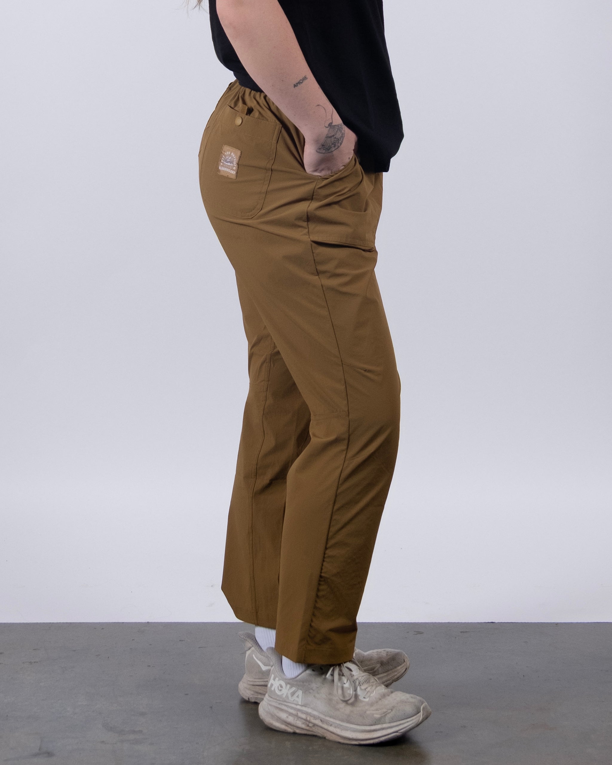 A person stands sideways against a plain light background wearing BEARDEDGOAT's NEW — Summit™ Pant in brown, paired with a black shirt, white socks, and sneakers. Their left hand is in their pocket, displaying a wrist tattoo.