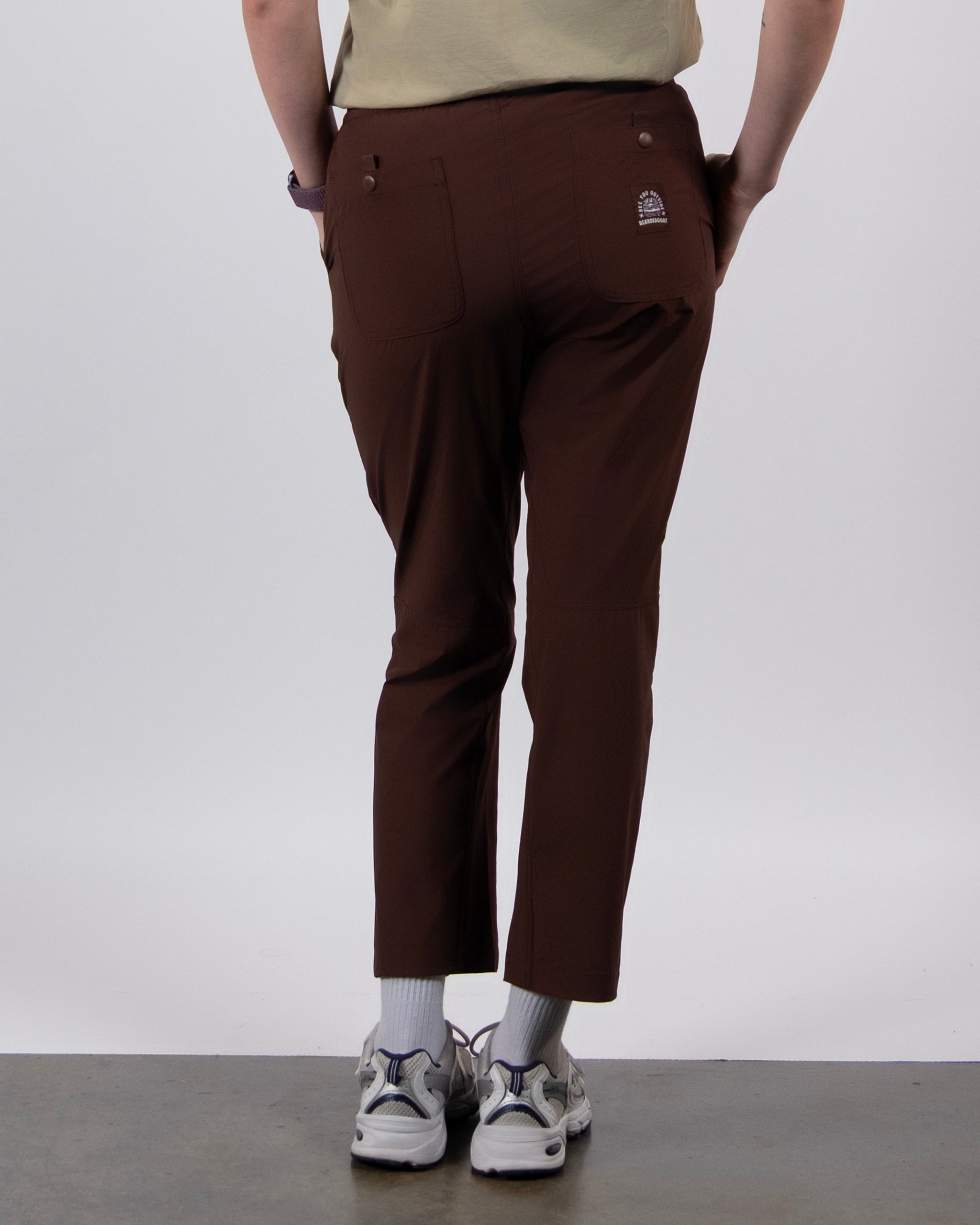 A person stands facing away, wearing BEARDEDGOAT's NEW — Summit™ Pant in brown with a light beige shirt, white socks, and white sneakers, hands tucked in the back pockets, against a plain light background.