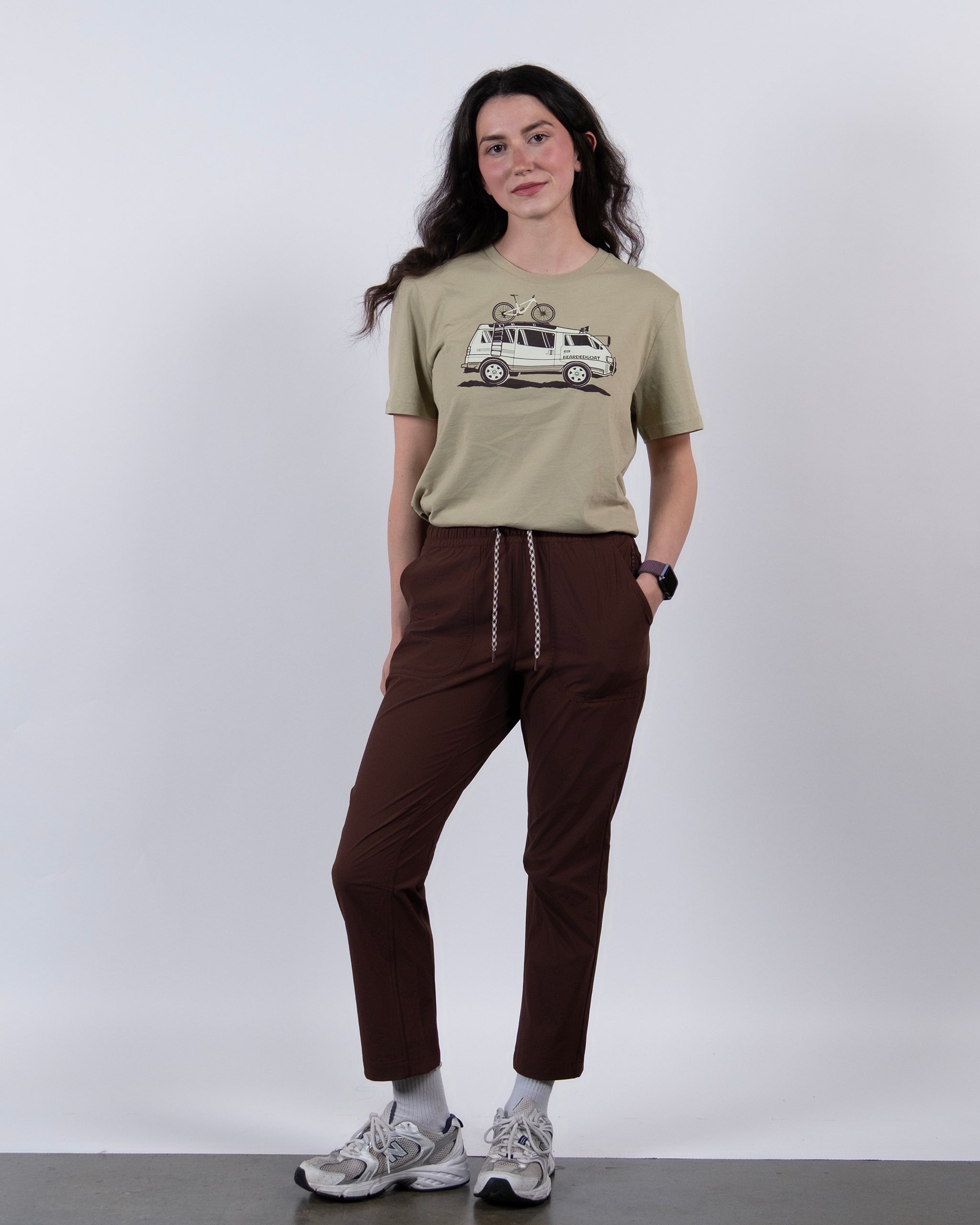 A woman with long dark hair stands against a plain background, wearing a beige graphic tee, BEARDEDGOAT’s NEW — Summit™ Pant with a drawstring, white sneakers, and a smartwatch.