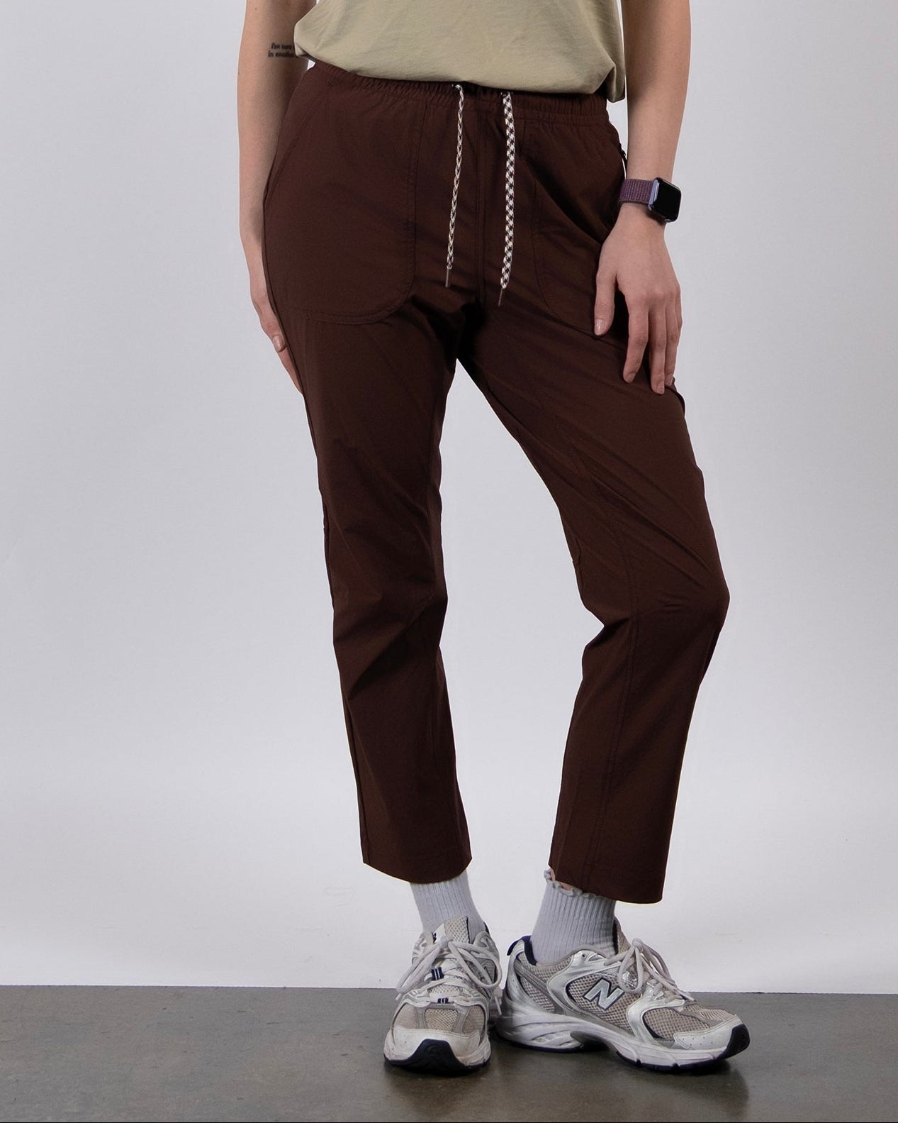 A woman with long brown hair stands against a white background, wearing a beige graphic T-shirt, BEARDEDGOAT NEW — Summit™ Pant in brown, gray socks, and white sneakers.