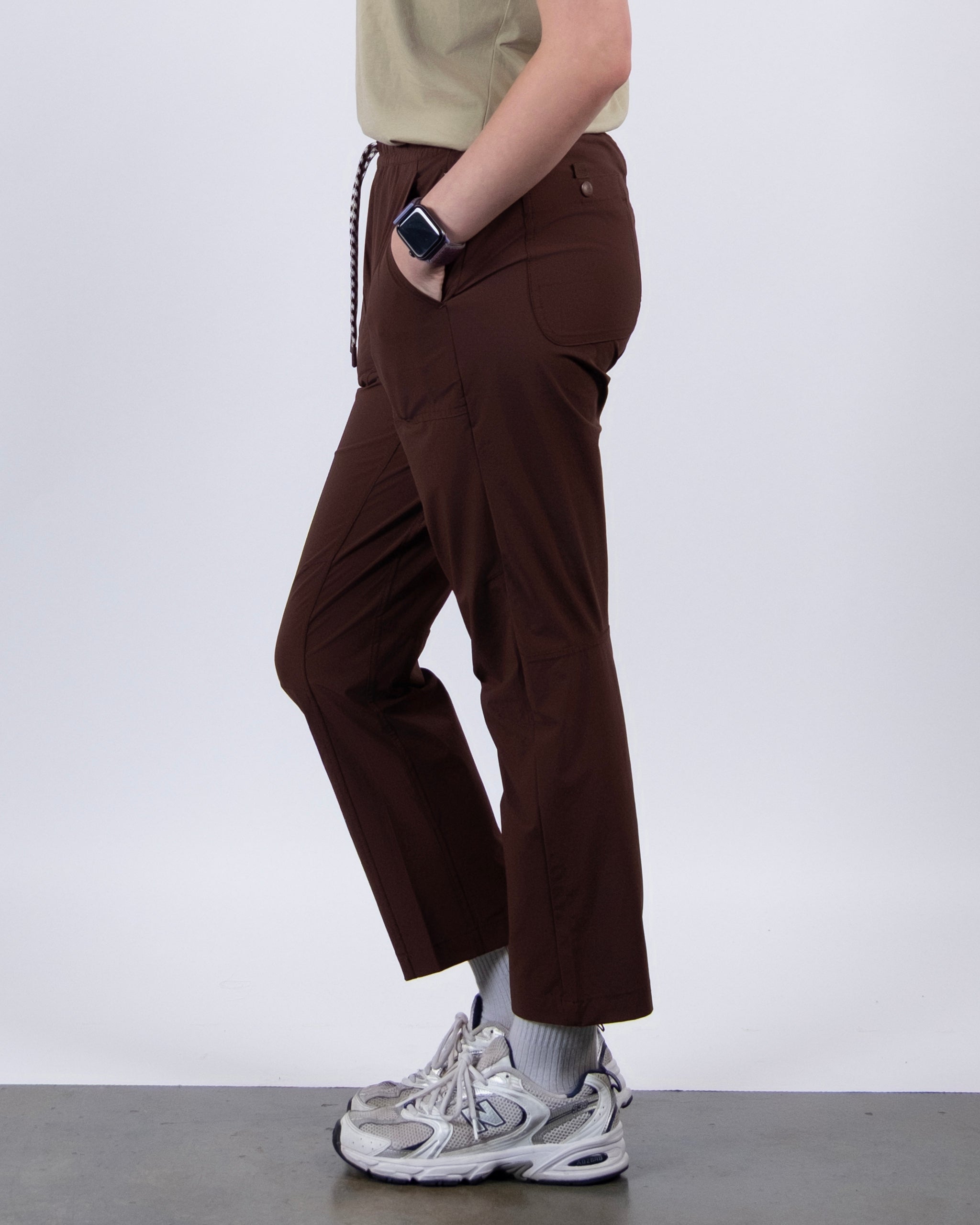 A person stands sideways against a plain light background wearing BEARDEDGOAT NEW — Summit™ Pant in brown, a beige shirt, white socks, gray sneakers, and a smartwatch.