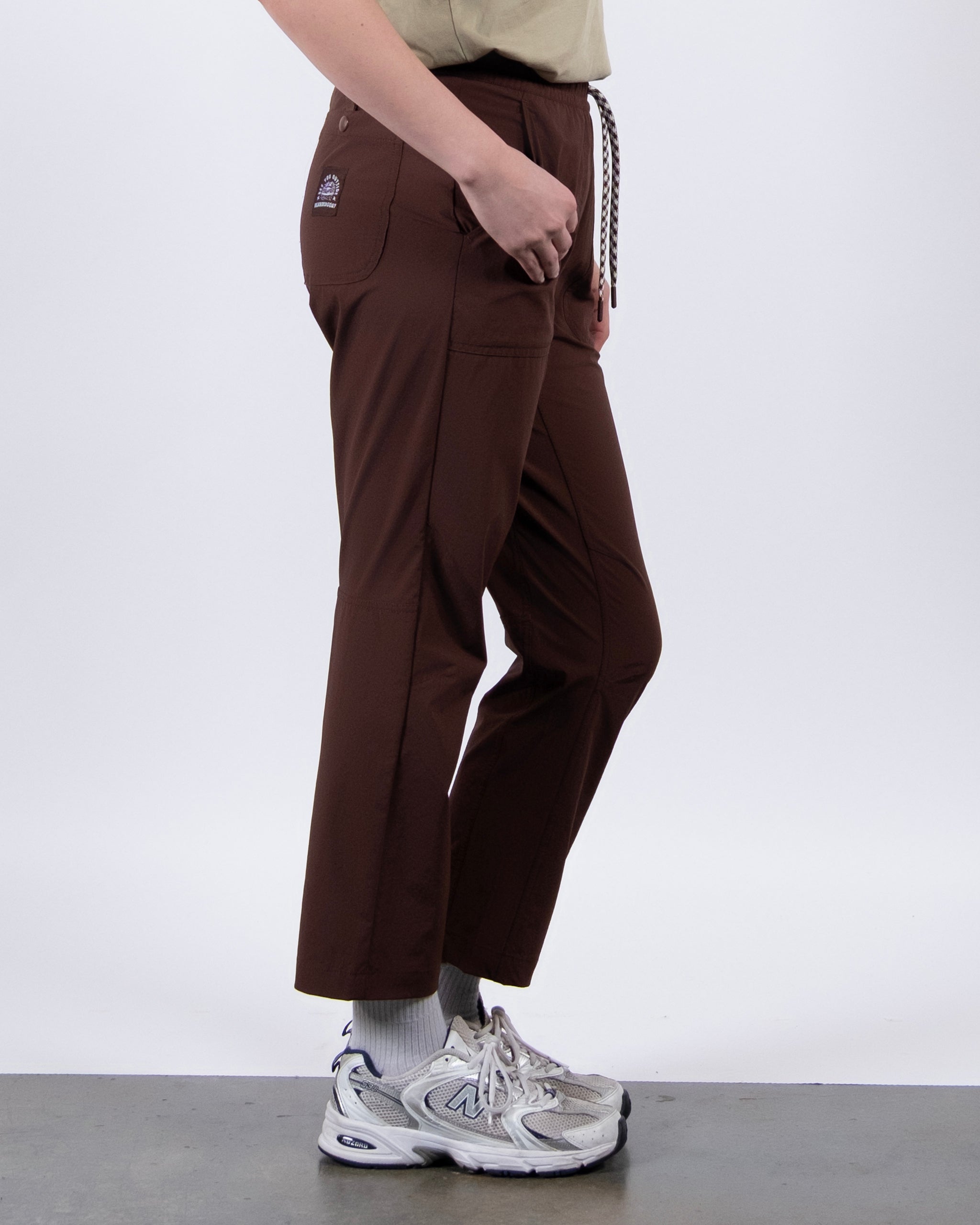 A person stands sideways in BEARDEDGOAT's NEW — Summit™ Pant, paired with a beige shirt, white socks, and gray sneakers. Their left hand is in the front pocket of the pants against a plain background.