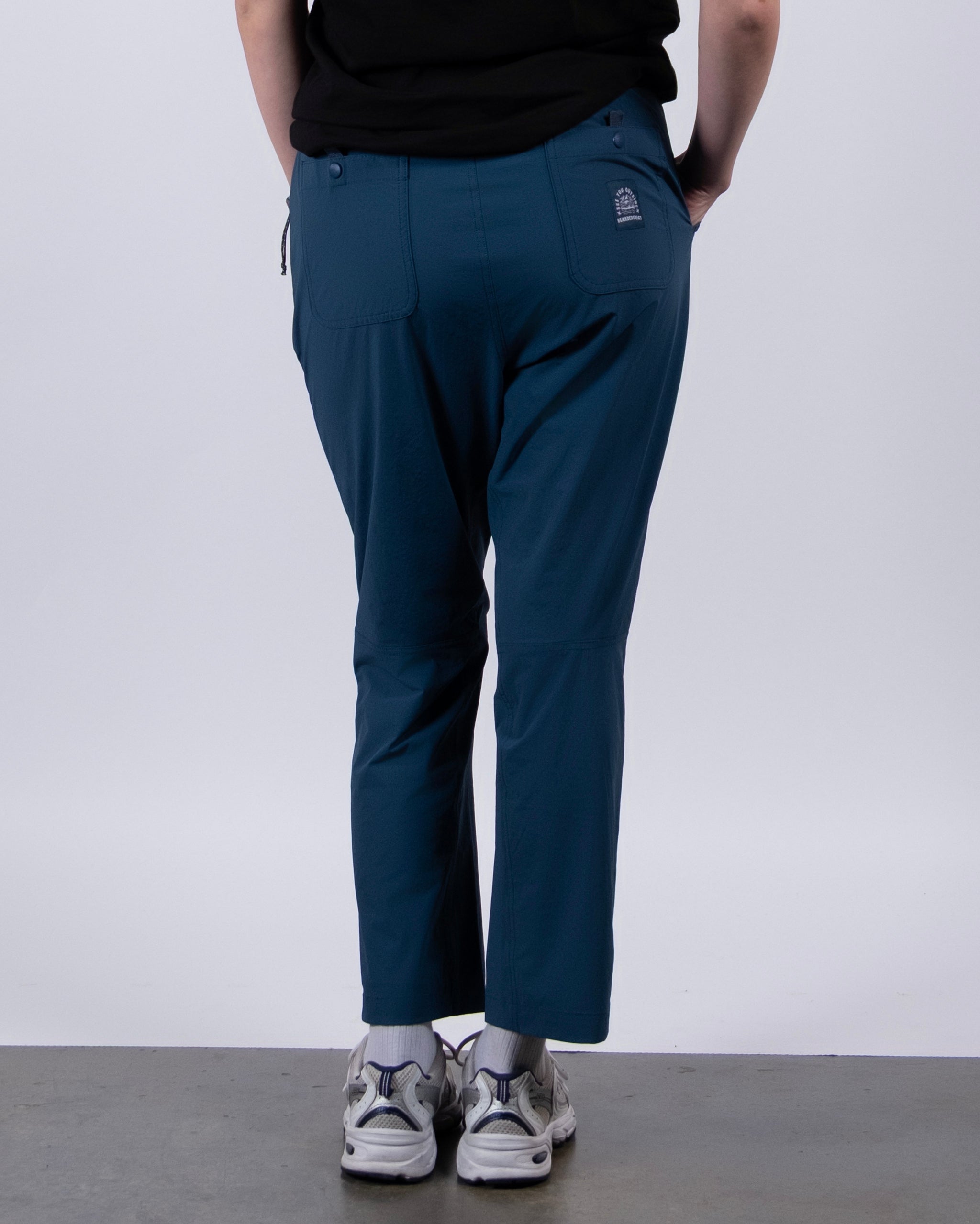 A person stands facing away from the camera, wearing a black shirt, white sneakers, and BEARDEDGOAT’s NEW — Summit™ Pant in teal blue against a plain light background.