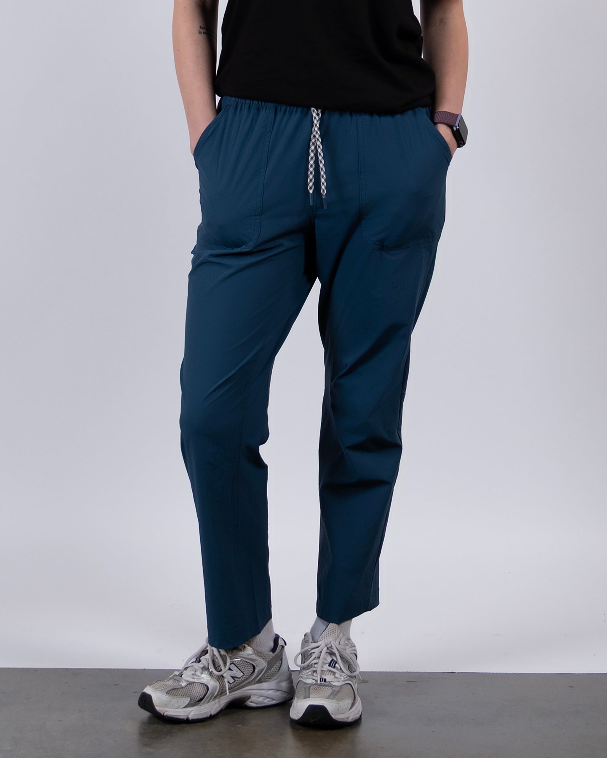 Wearing BEARDEDGOAT’s NEW — Summit™ Pant in teal, paired with a black shirt, gray running shoes, and a smartwatch, a woman stands with hands in pockets against a simple light background.
