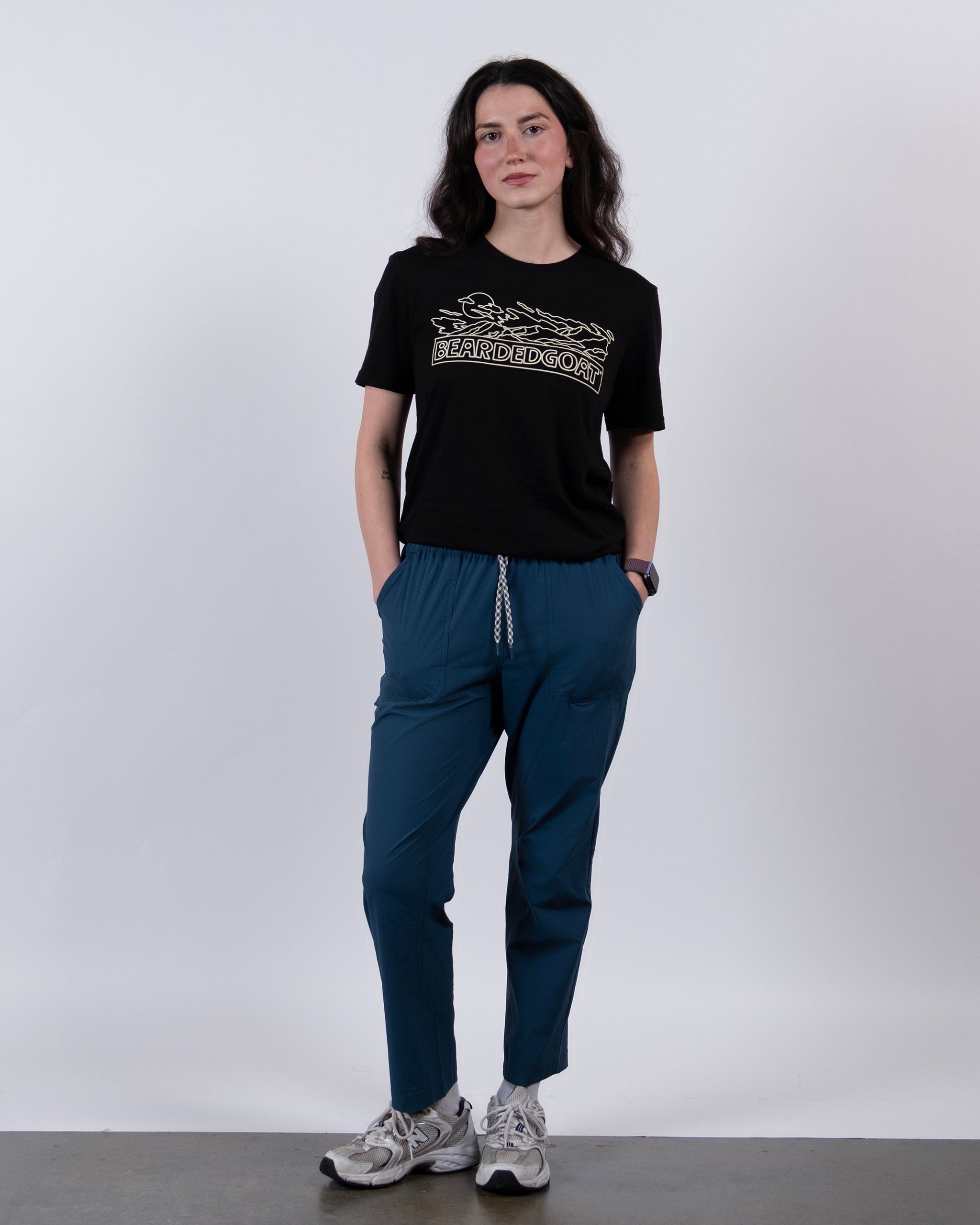 A woman with long dark hair stands against a plain background, wearing a black graphic T-shirt, BEARDEDGOAT's NEW — Summit™ Pant in teal, and gray sneakers. Her hands are in her pockets and she has a neutral expression.