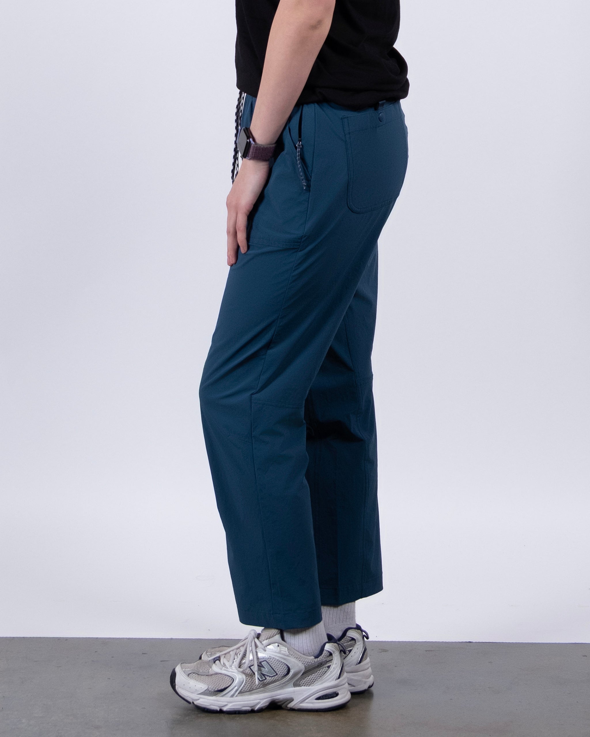 A person stands sideways against a plain light background, wearing BEARDEDGOAT’s NEW — Summit™ Pant in teal (stain-resistant), a black shirt, white socks, gray athletic sneakers, and a watch.