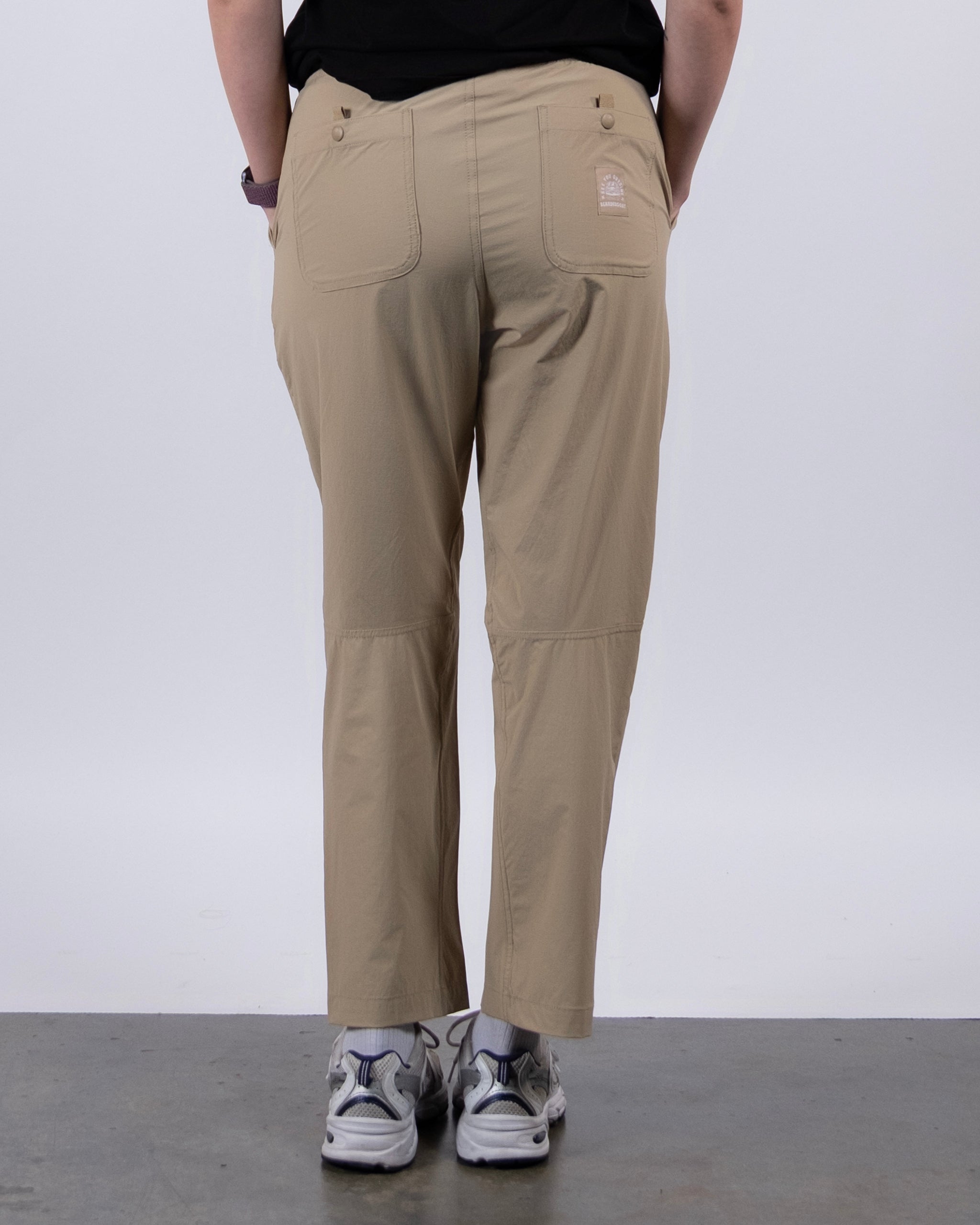 Someone is wearing BEARDEDGOAT's NEW — Summit™ Pant in beige and gray sneakers, standing on a gray floor with their back to the camera against a plain light background.