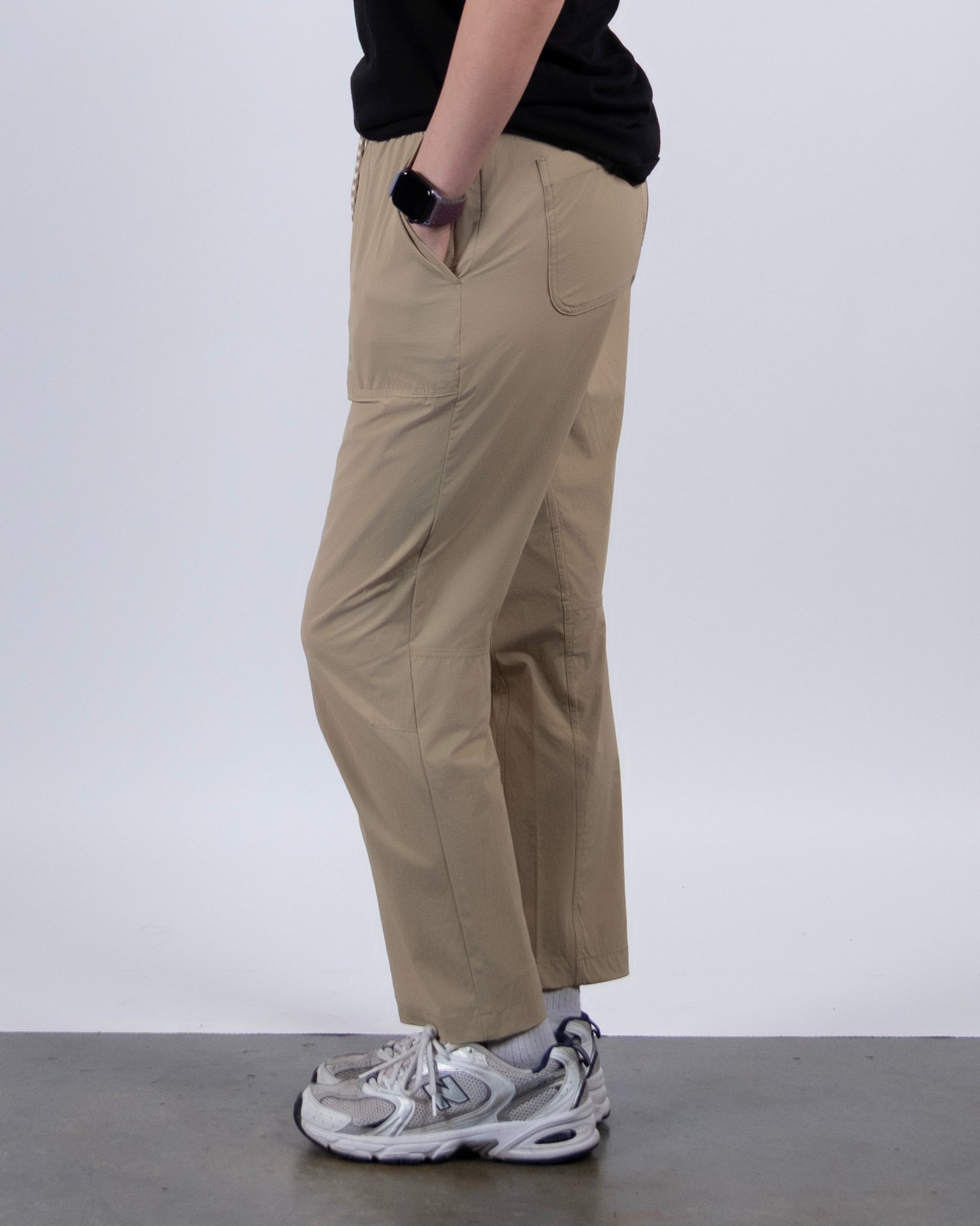A person stands sideways with one hand in their pocket, wearing BEARDEDGOAT’s NEW — Summit™ Pant in beige, a black shirt, white socks, and silver athletic shoes against a plain background.