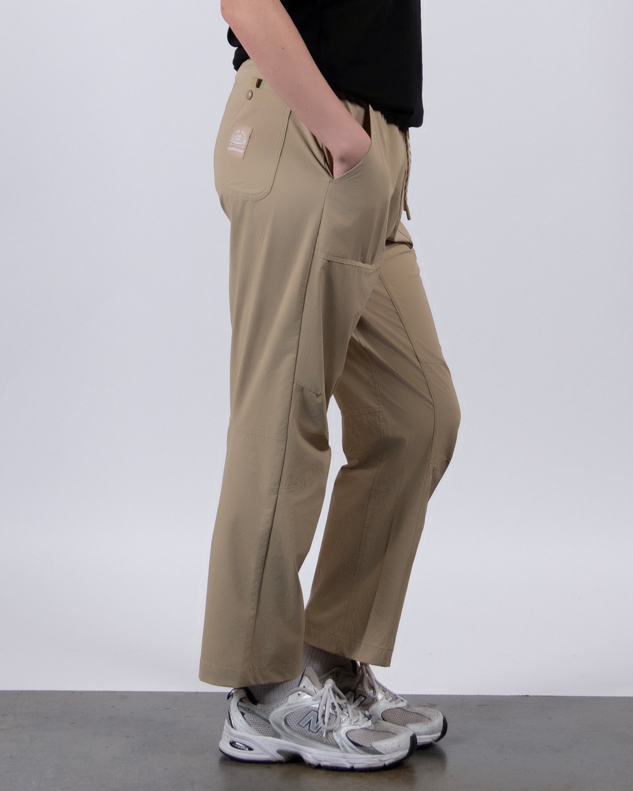 A person stands sideways against a white background, wearing BEARDEDGOAT NEW — Summit™ Pant in light brown, paired with a black top and gray athletic shoes, one hand in their pocket.