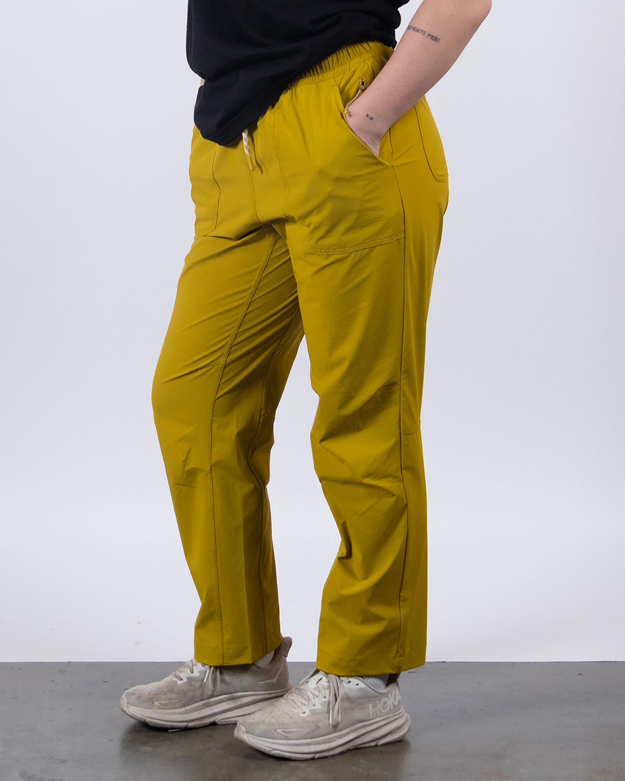 A person models the BEARDEDGOAT NEW — Summit™ Pant in mustard yellow, paired with a black shirt and white athletic shoes. Hands are in pockets, only the lower half of the body is shown against a plain white background.
