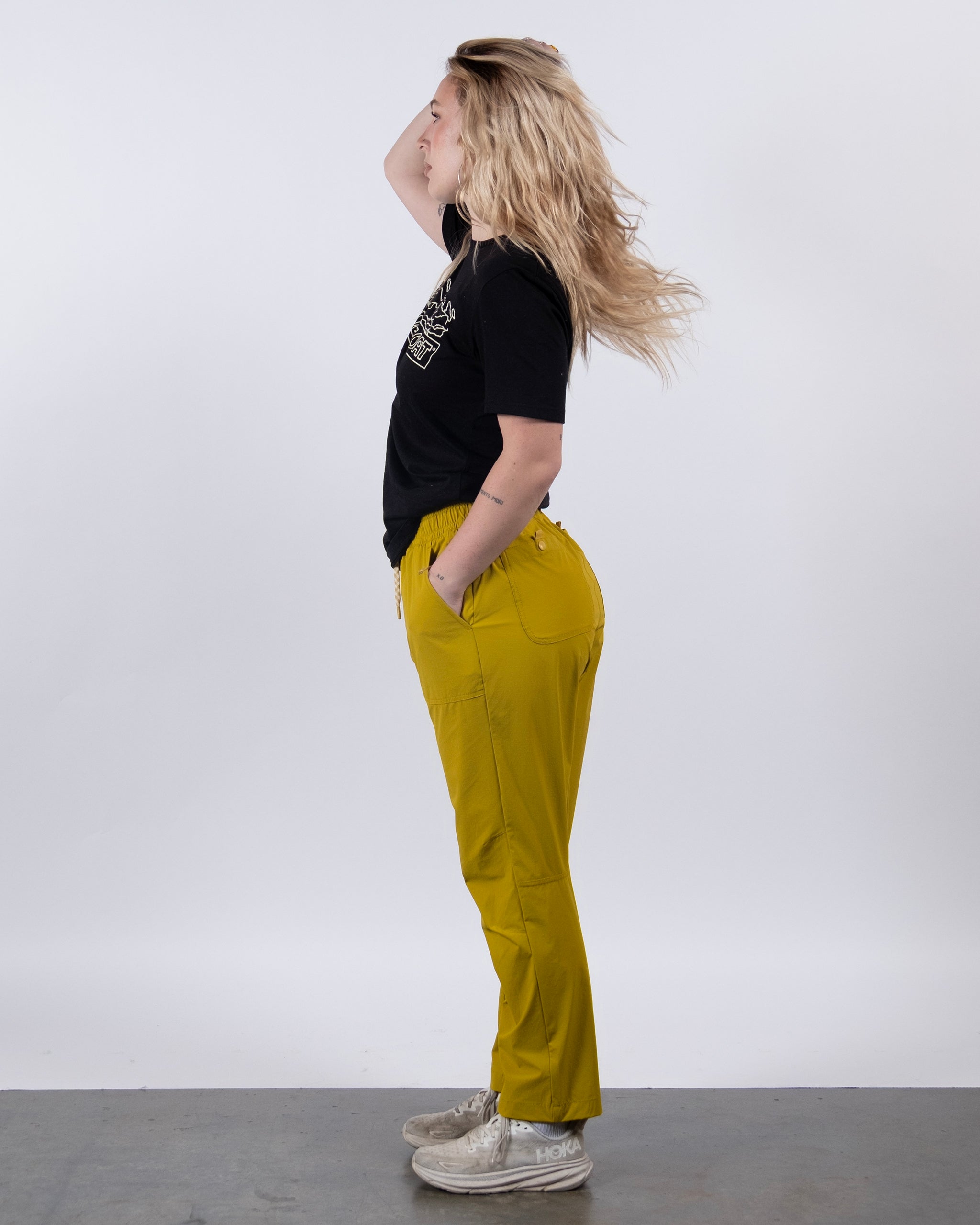 A person with long blonde hair stands in profile against a white background, wearing a black T-shirt, BEARDEDGOAT NEW — Summit™ Pant in yellow, and white sneakers, with one hand on their head and the other in their pocket.