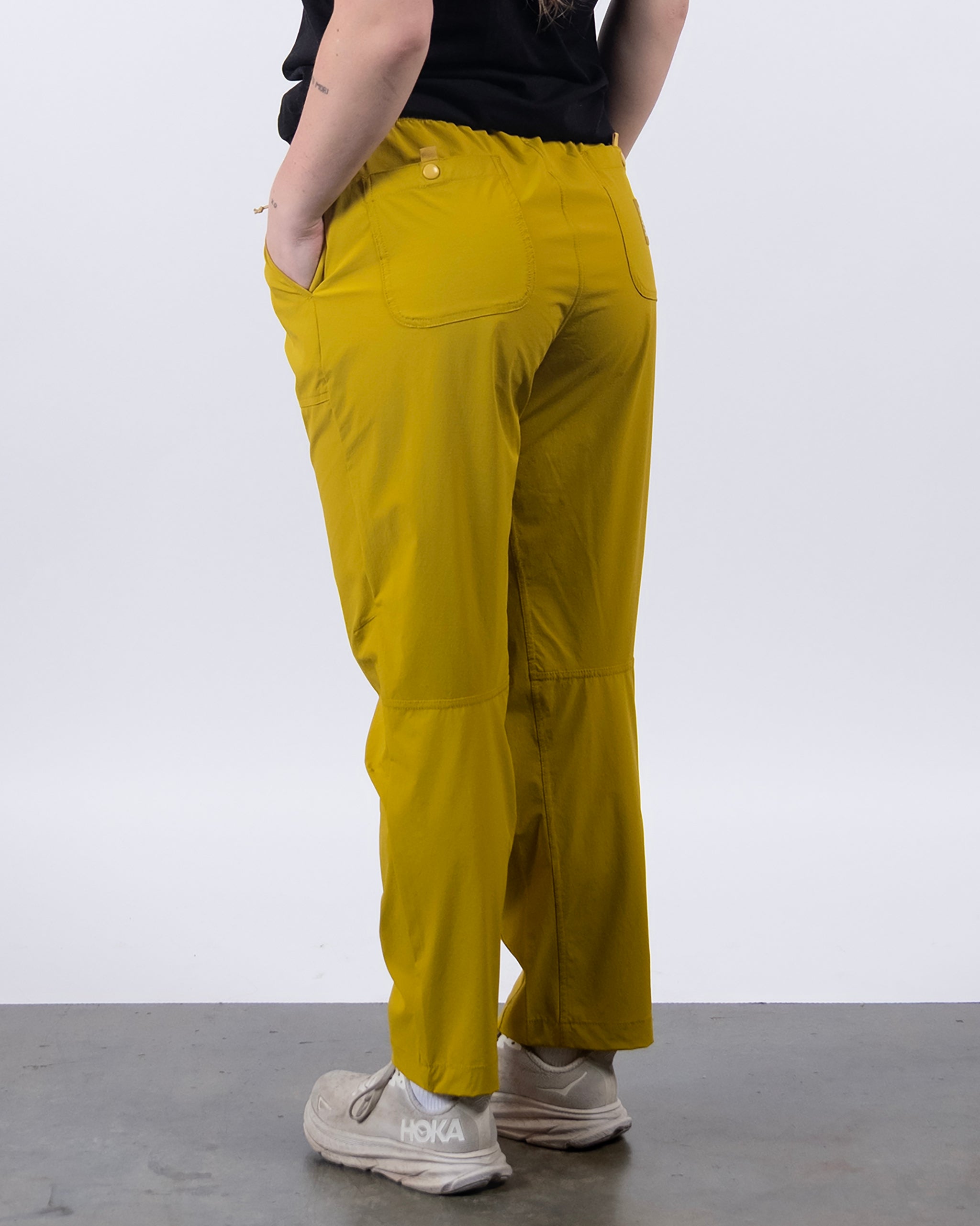 A person models BEARDEDGOAT's NEW — Summit™ Pant in mustard yellow, featuring back pockets, stain-resistant and water-repellent fabric, paired with white Hoka sneakers against a plain white background, hands in pockets, facing away.