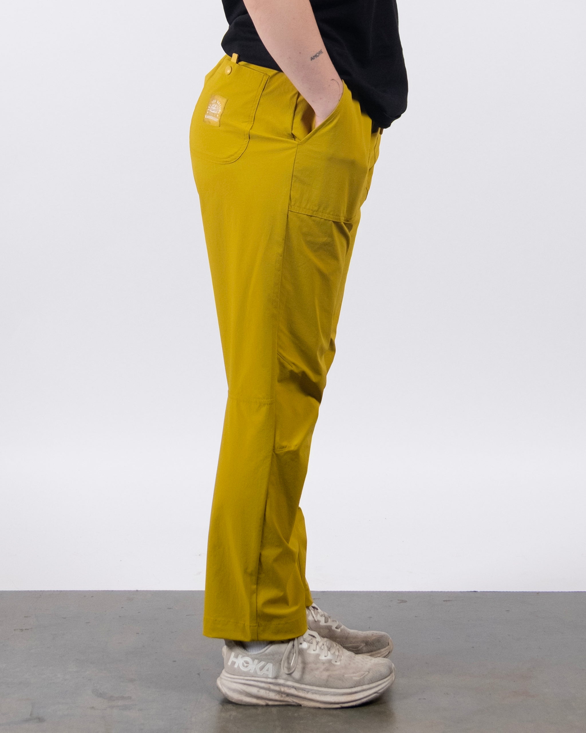 A person stands sideways with only their lower half visible, wearing BEARDEDGOAT’s NEW — Summit™ Pant in mustard yellow, paired with a black shirt and white athletic shoes against a plain, light background.