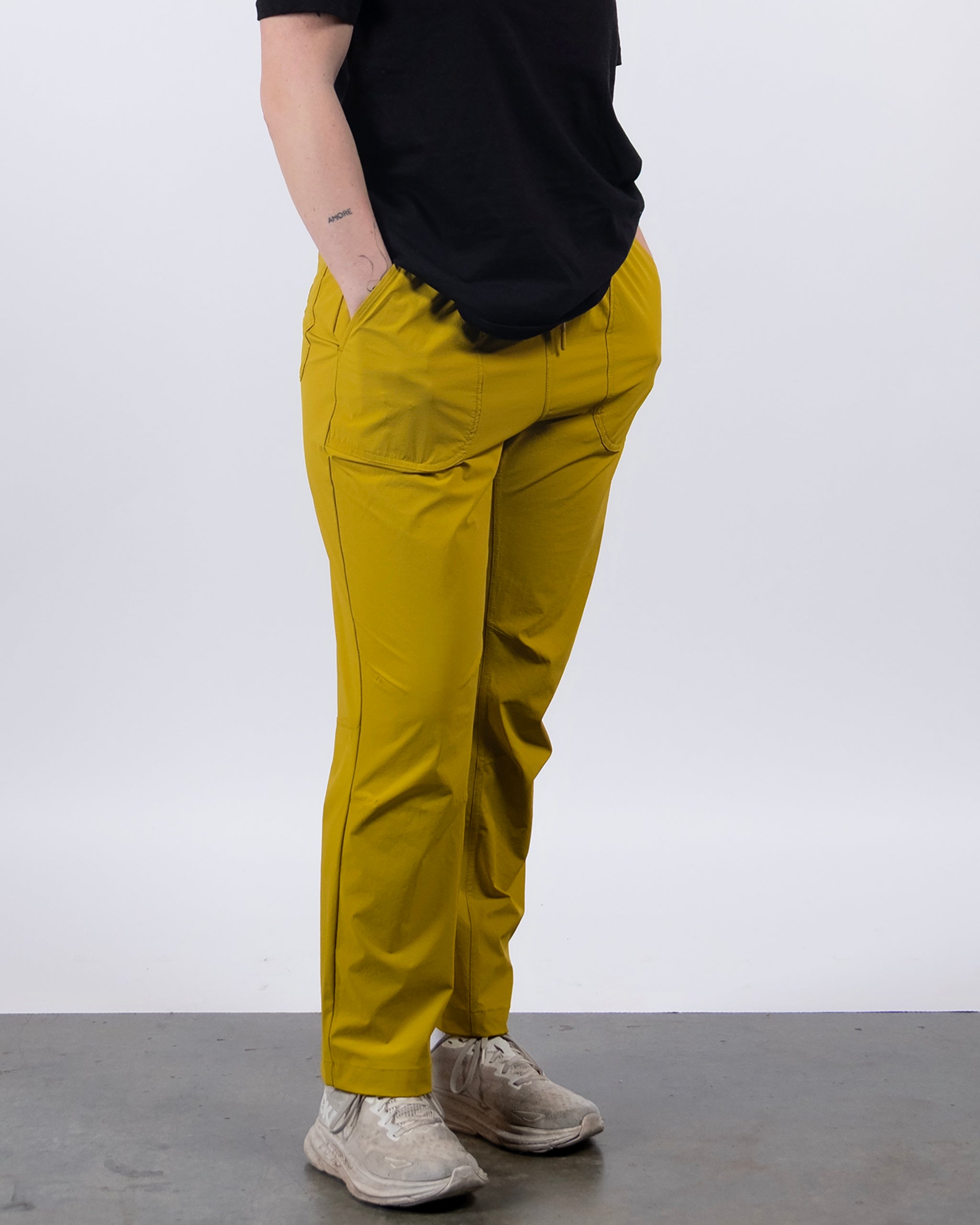 A person wearing a black shirt, BEARDEDGOAT NEW — Summit™ Pant in mustard yellow, and light-colored sneakers stands with hands in pockets against a plain white background.