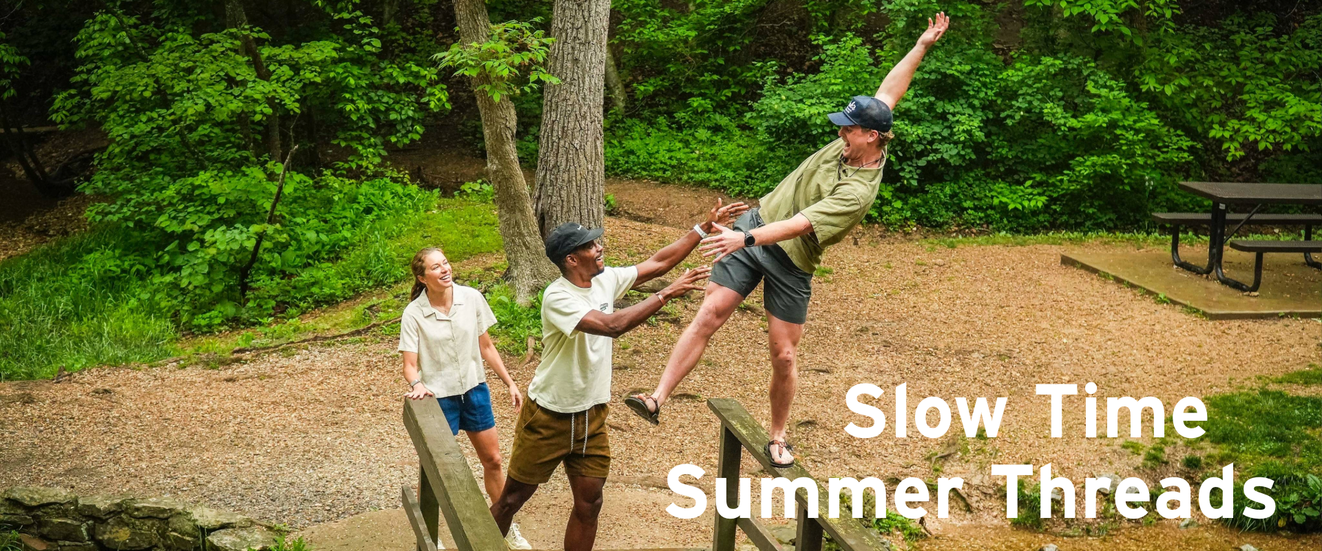 Three friends enjoy a sunny day in a wooded park; two help the third balance on a wooden rail. A picnic table is in the background. Text reads: Slow Time Summer Threads.