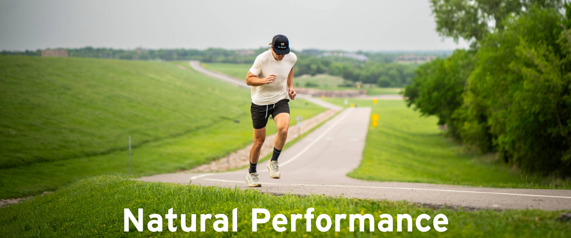 A man in athletic wear runs uphill on a paved path through green fields, with trees and open sky in the background. The text Natural Performance appears at the bottom of the image.