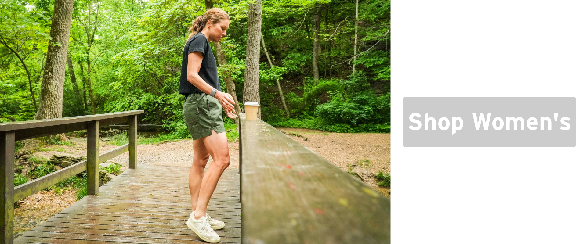 A person in a black shirt and green shorts stands on a wooden bridge in a lush, green forest, holding a drink. Text on the right reads: Shop Womens.