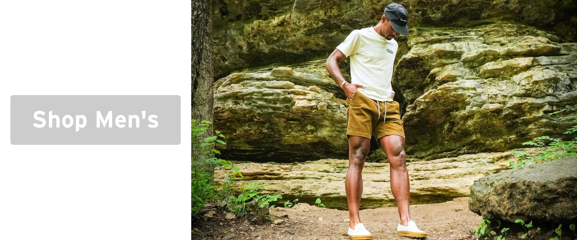 A man wearing a white t-shirt, brown shorts, white shoes, and a black cap stands outdoors on a dirt path with rock formations behind him. To the left, a button reads Shop Mens.