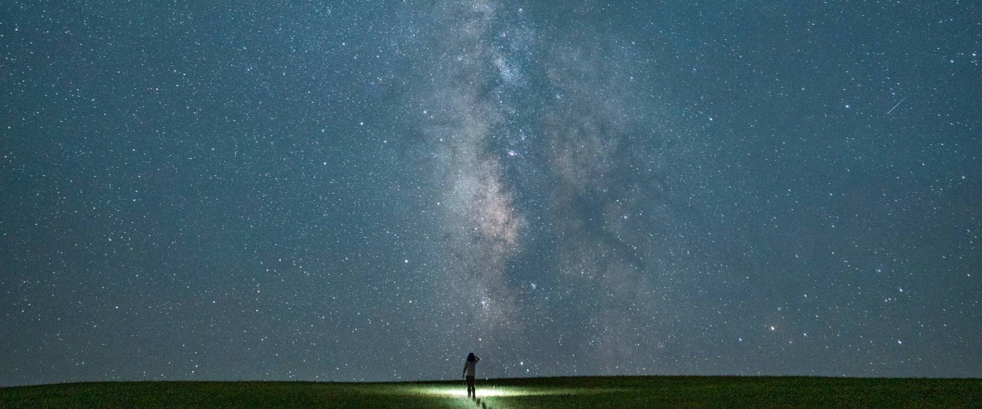A person stands on a grassy hill at night, illuminated by a flashlight, gazing up at a bright, star-filled sky dominated by the Milky Way galaxy.