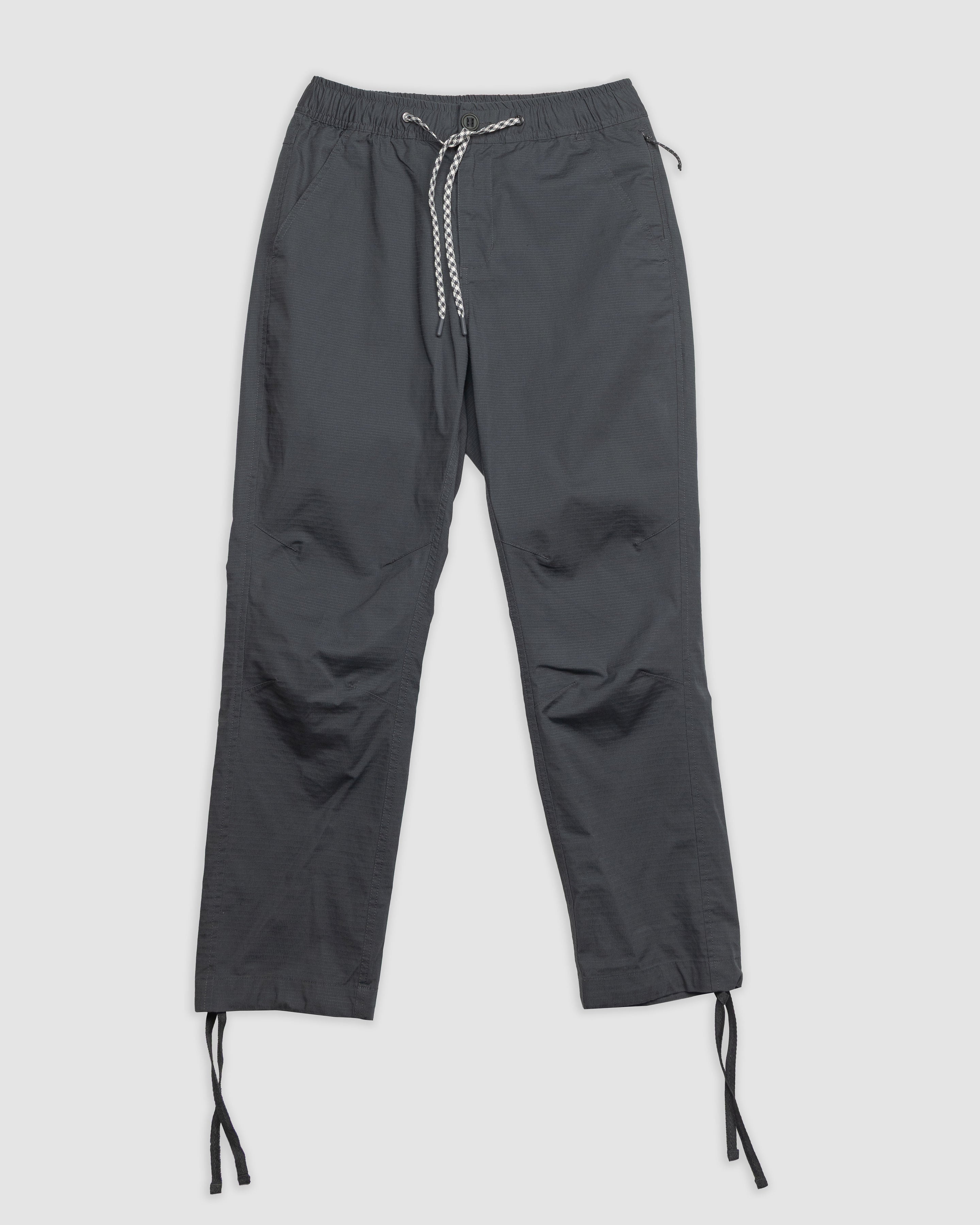 The Womens Canopy Pant by BEARDEDGOAT in dark gray features stretch cotton ripstop, an elastic waistband, side pockets, and adjustable ankle drawcords. Shown laid flat on a light background.