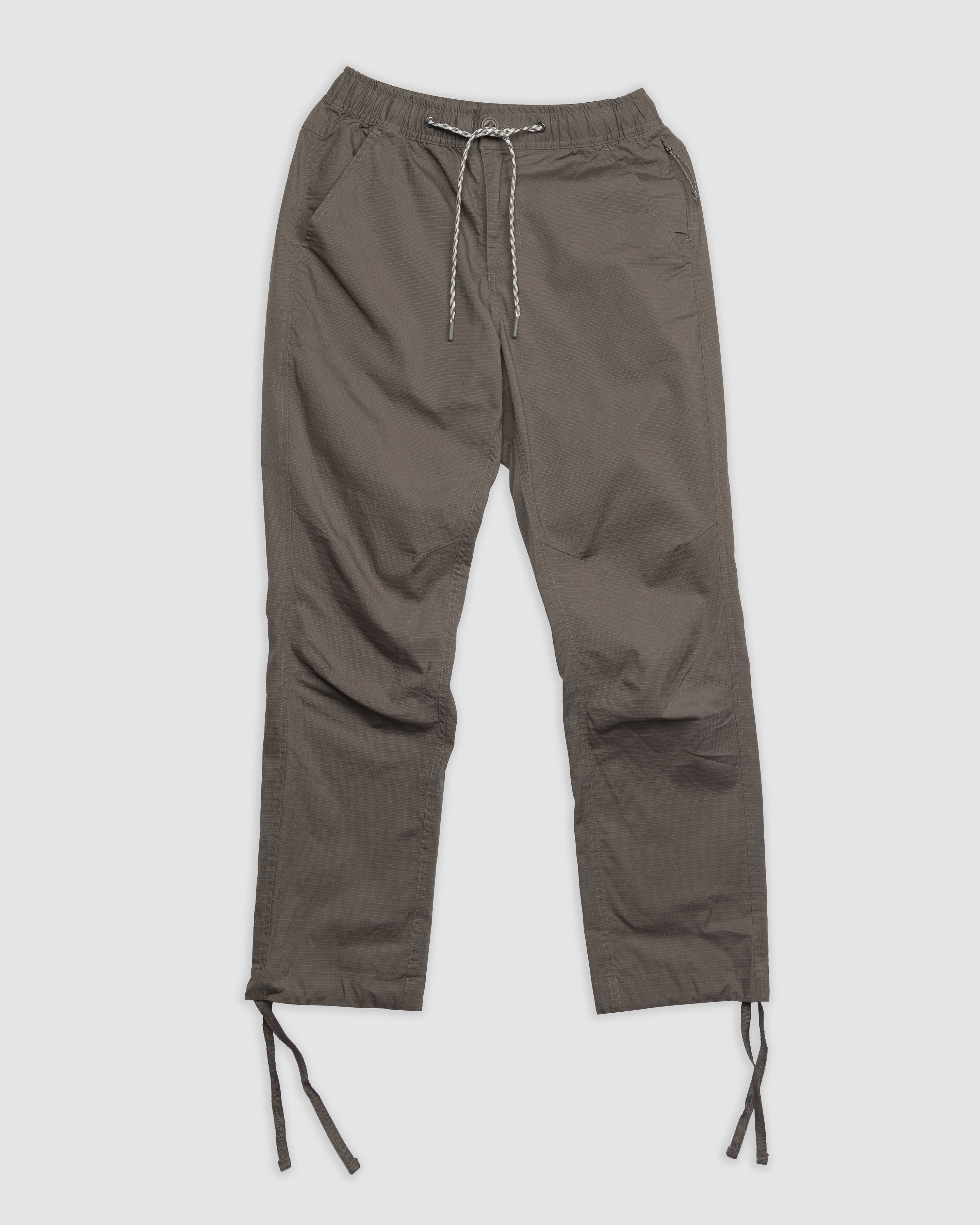 The Womens Canopy Pant by BEARDEDGOAT in olive green, made from stretch cotton ripstop with side pockets, elastic waistband, and adjustable ankle drawcords, is displayed flat on a light gray background.