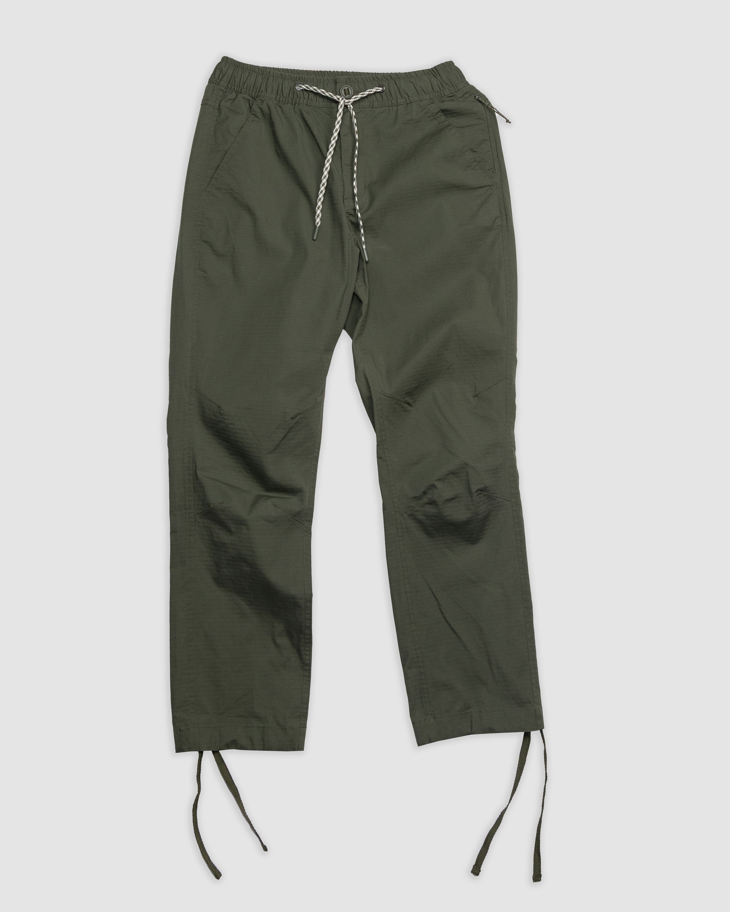 The BEARDEDGOAT Womens Canopy Pant in olive green, made from stretch cotton ripstop, features an elastic waistband, two side pockets, and adjustable ankle drawstrings. Shown laid flat on a white background.