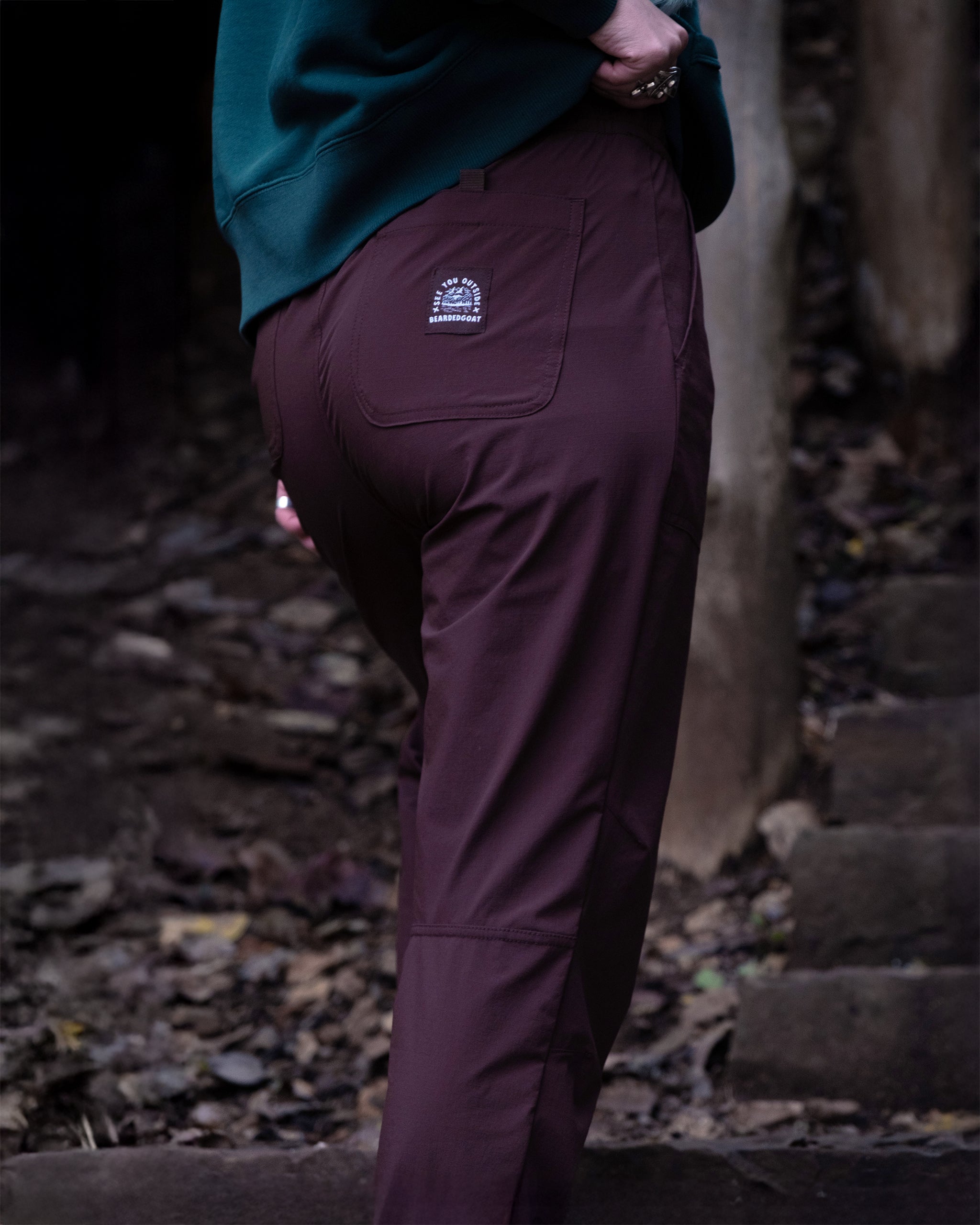Wearing BEARDEDGOAT’s new Summit™ Pant in dark brown and a green sweatshirt, a person stands on a leaf-covered path by stone steps. The durable stretch pants, ideal for outdoor adventures, have a logo patch on the back pocket.