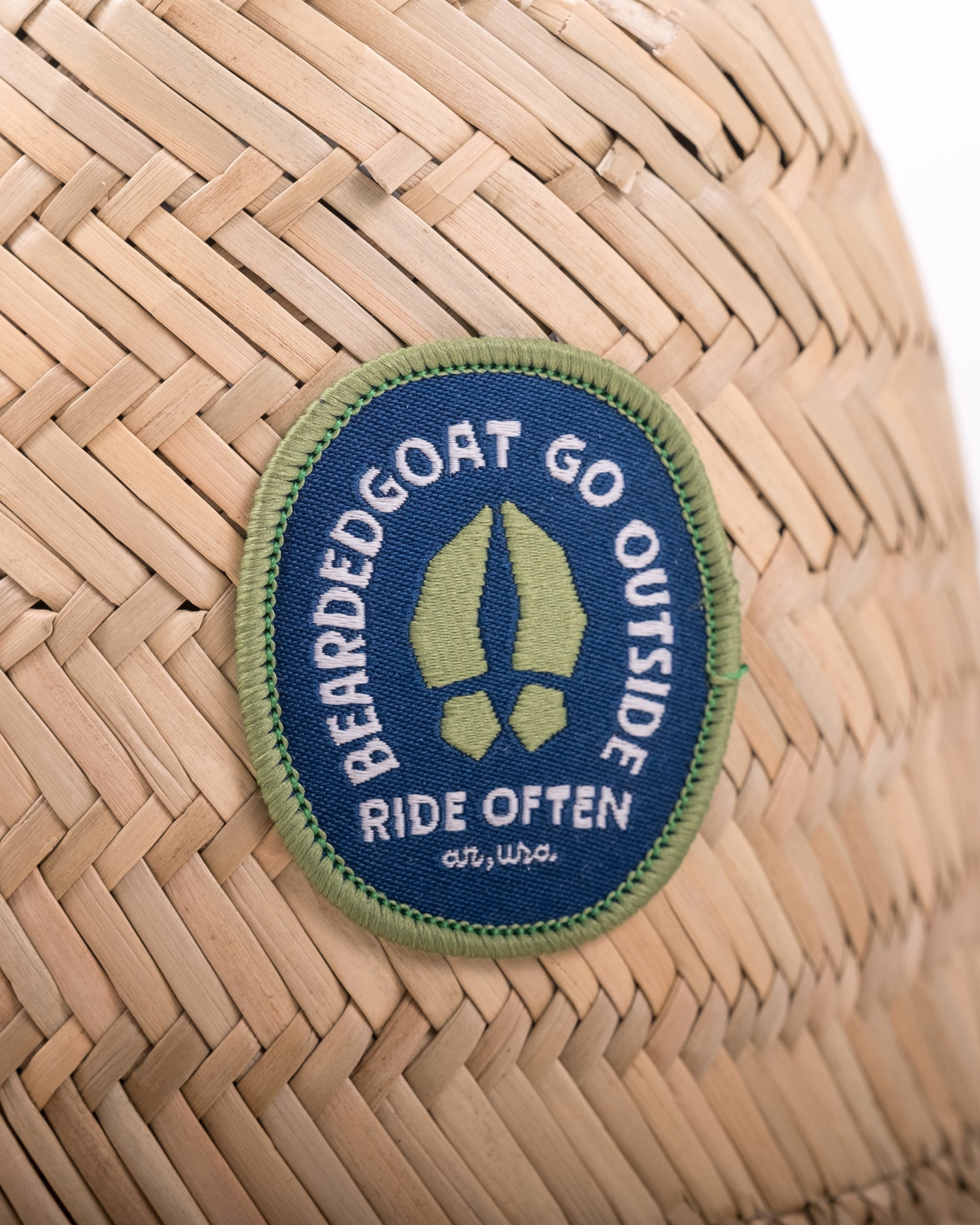 A close-up of the beardedgoat Sun Hat’s woven straw surface shows a blue and green patch reading BEARDEDGOAT GO OUTSIDE RIDE OFTEN ar, usa with a hoof print graphic, blending style with skin protection.