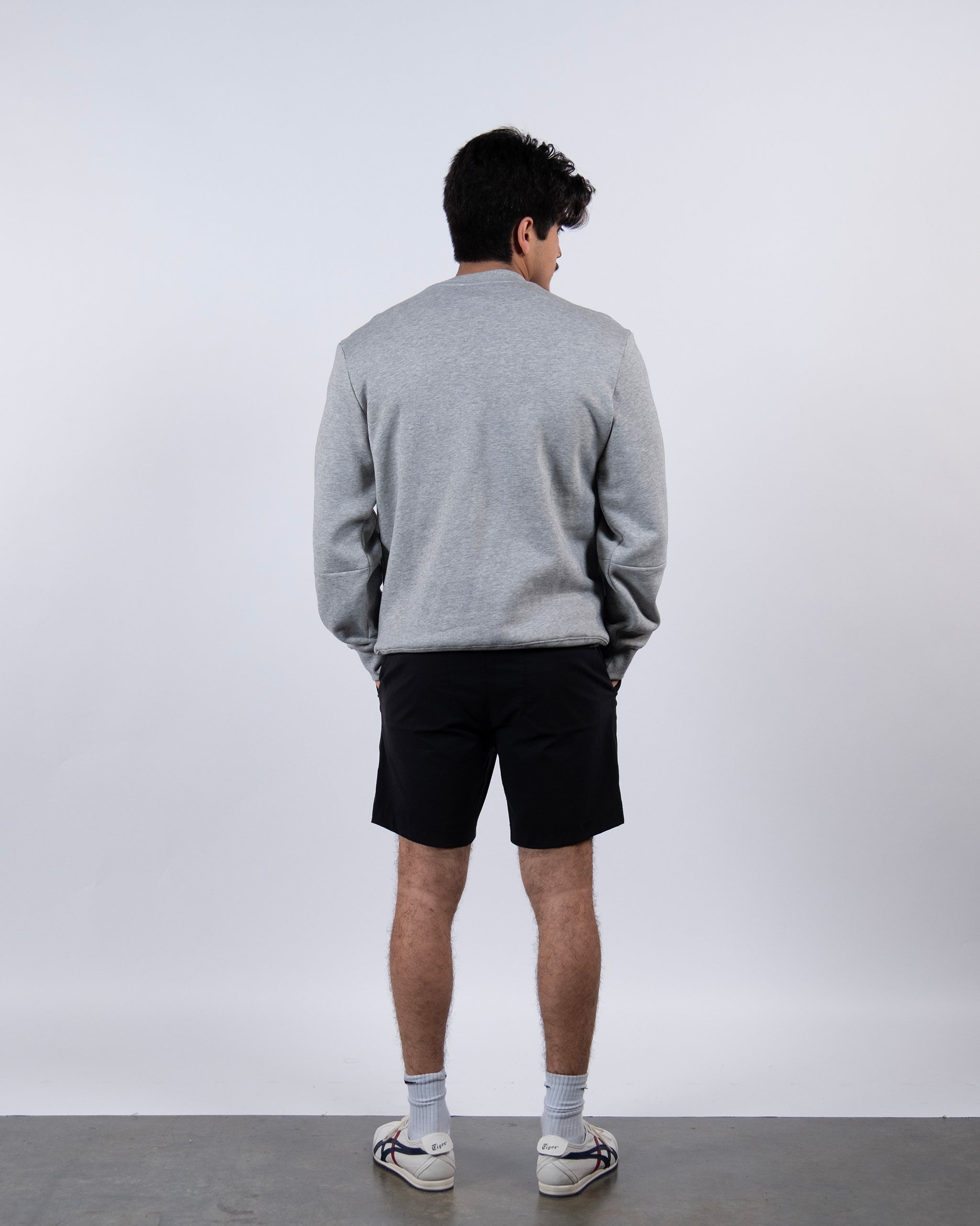 A person with short dark hair stands indoors facing away from the camera, wearing the beardedgoat Men's Void Crew in light gray heavyweight cotton fleece, black shorts, white socks, and white sneakers against a plain light background.