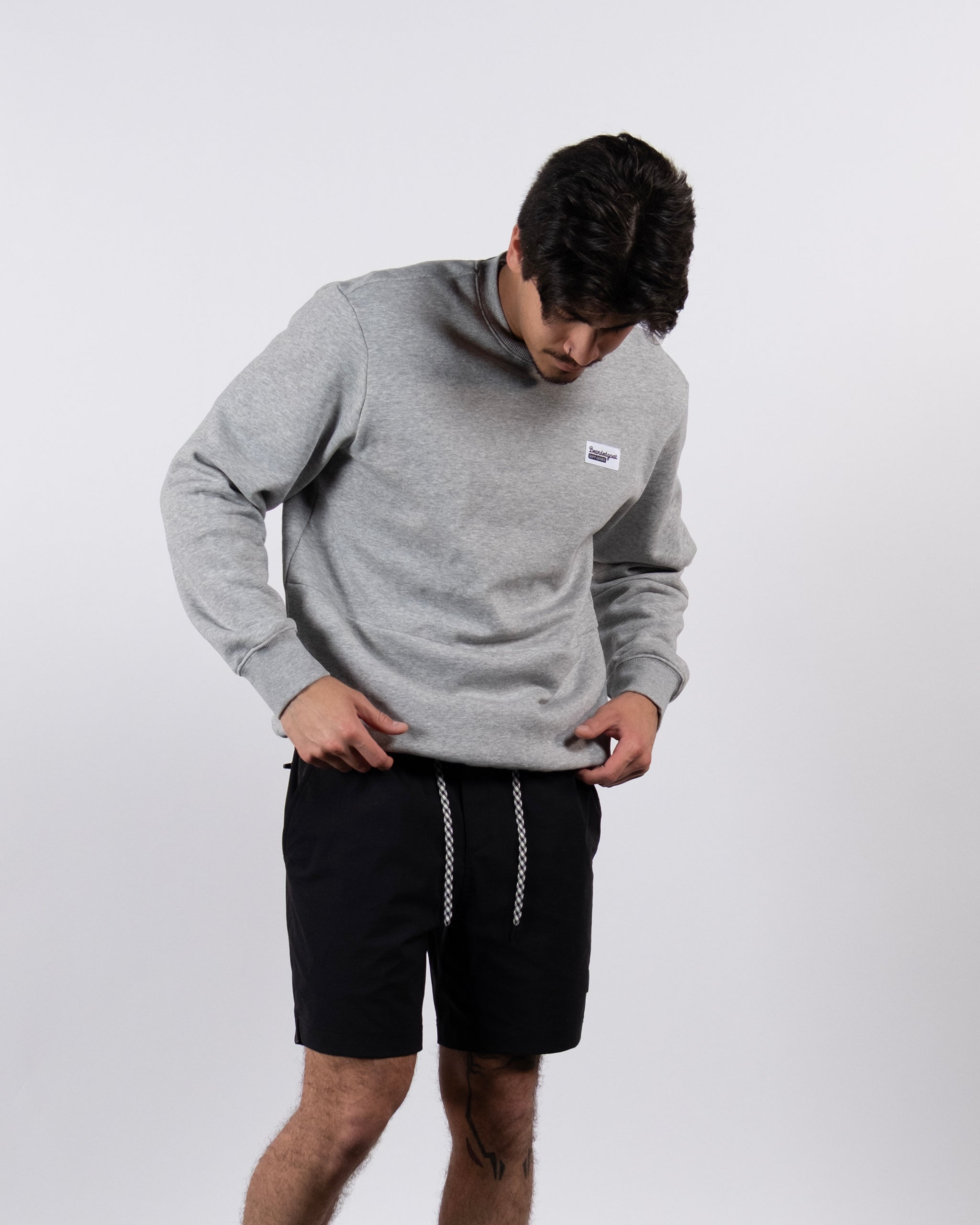 A man wearing the beardedgoat Men's Void Crew, a grey crewneck sweatshirt with ribbed knit cuffs, and black shorts stands against a plain white background, looking down as he adjusts his sweatshirt.